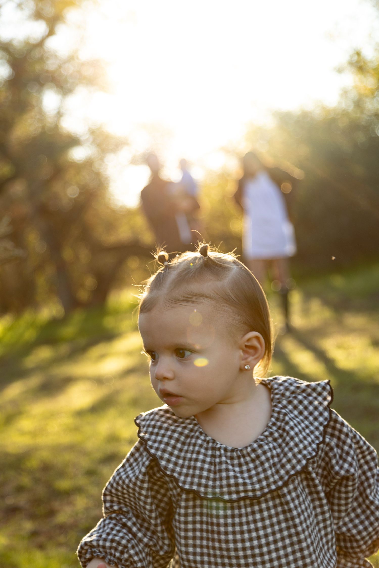 A toddler girl in a gingham dress explores a sunlit garden while her parents stand in the golden bokeh background.