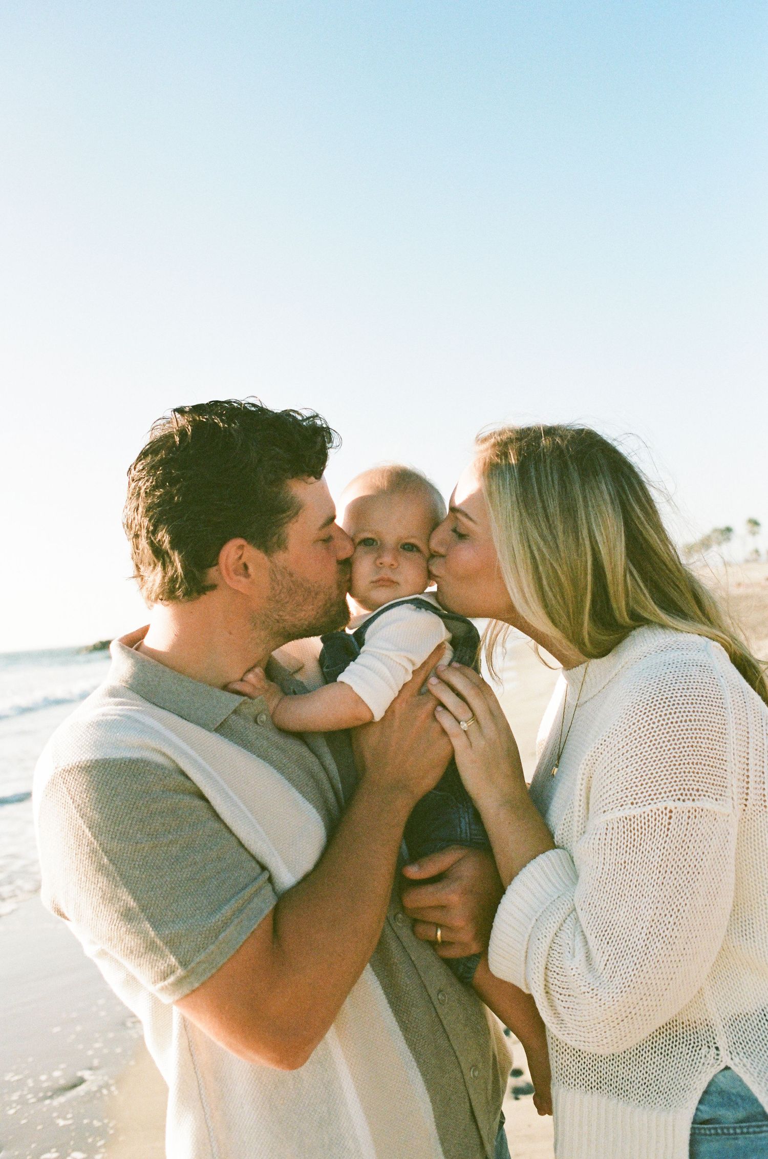 Parents kissing baby on cheeks on sunny beach, family portrait outdoors.