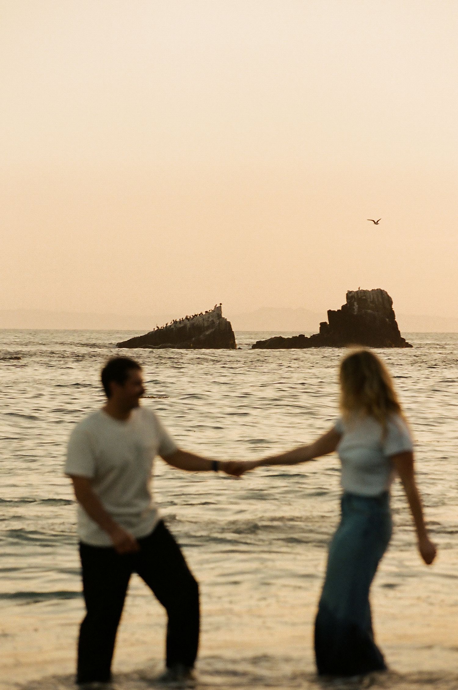 Couple holds hands in ocean water at warm sunset with silhouetted rocky island behind.