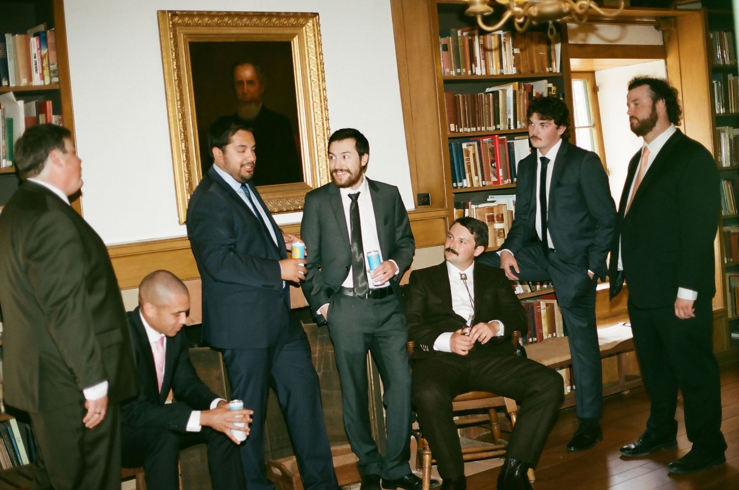 Group of men in dark suits socializing in an elegant library room with bookshelves and a portrait painting.