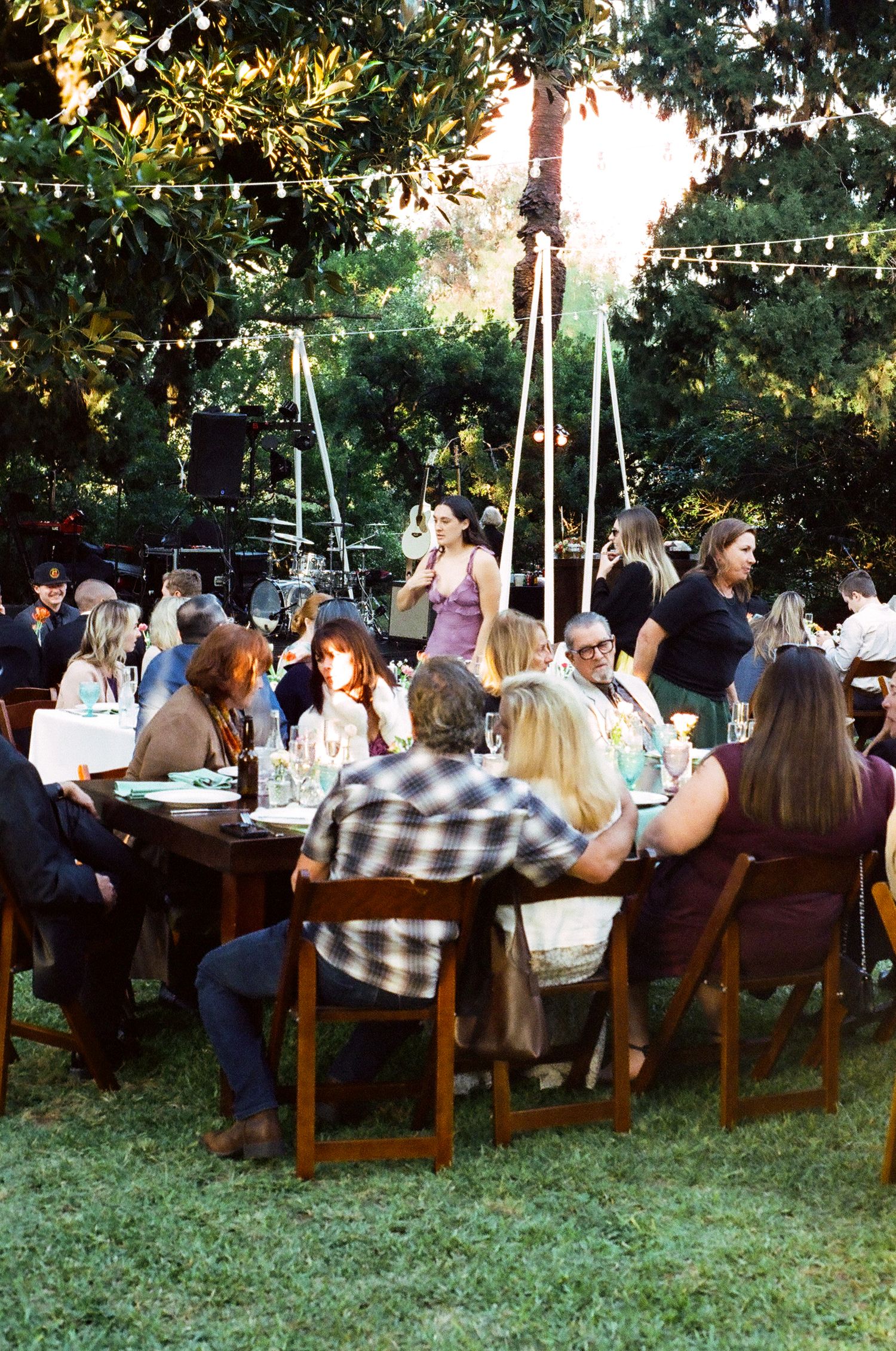 Guests gathered around rustic wooden tables at an outdoor garden dinner party with string lights and lush green trees.