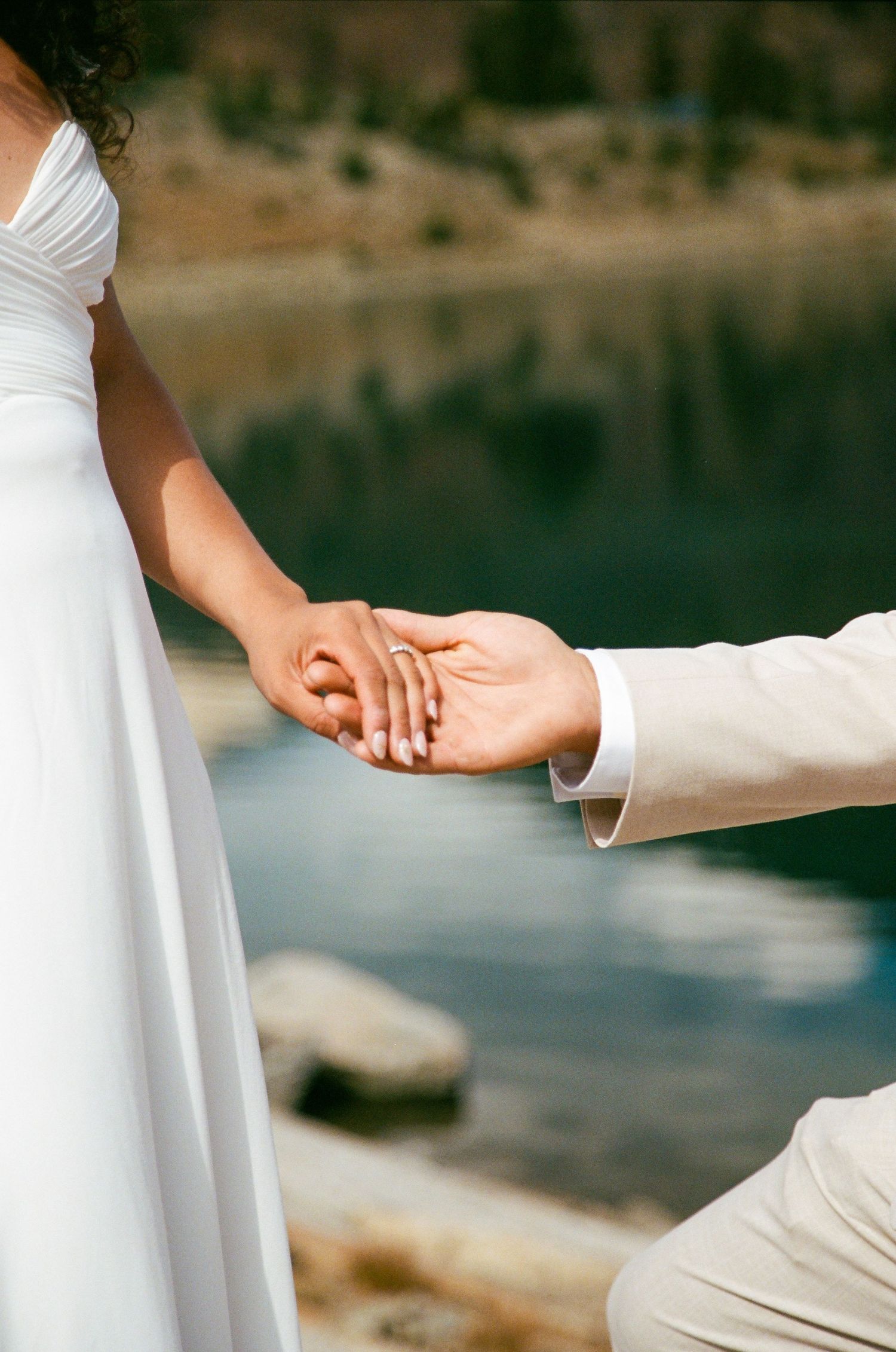 Bride and groom holding hands near a mountain lake, bride wearing white gown with diamond ring visible.