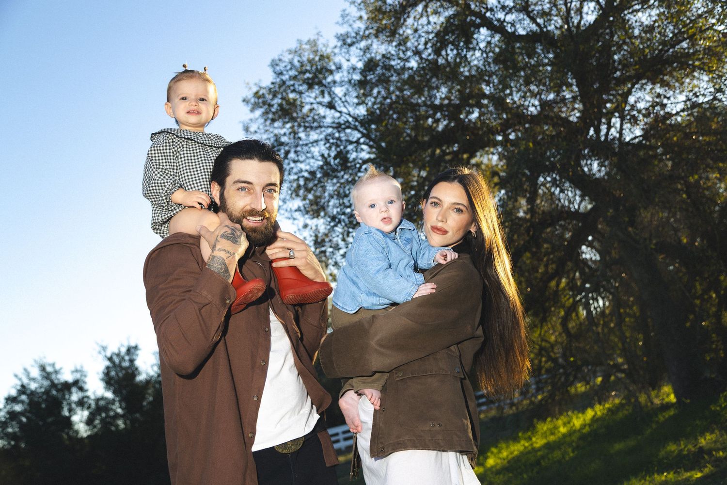 Smiling family of four poses outdoors, parents each holding a baby under a large tree in warm sunlight.