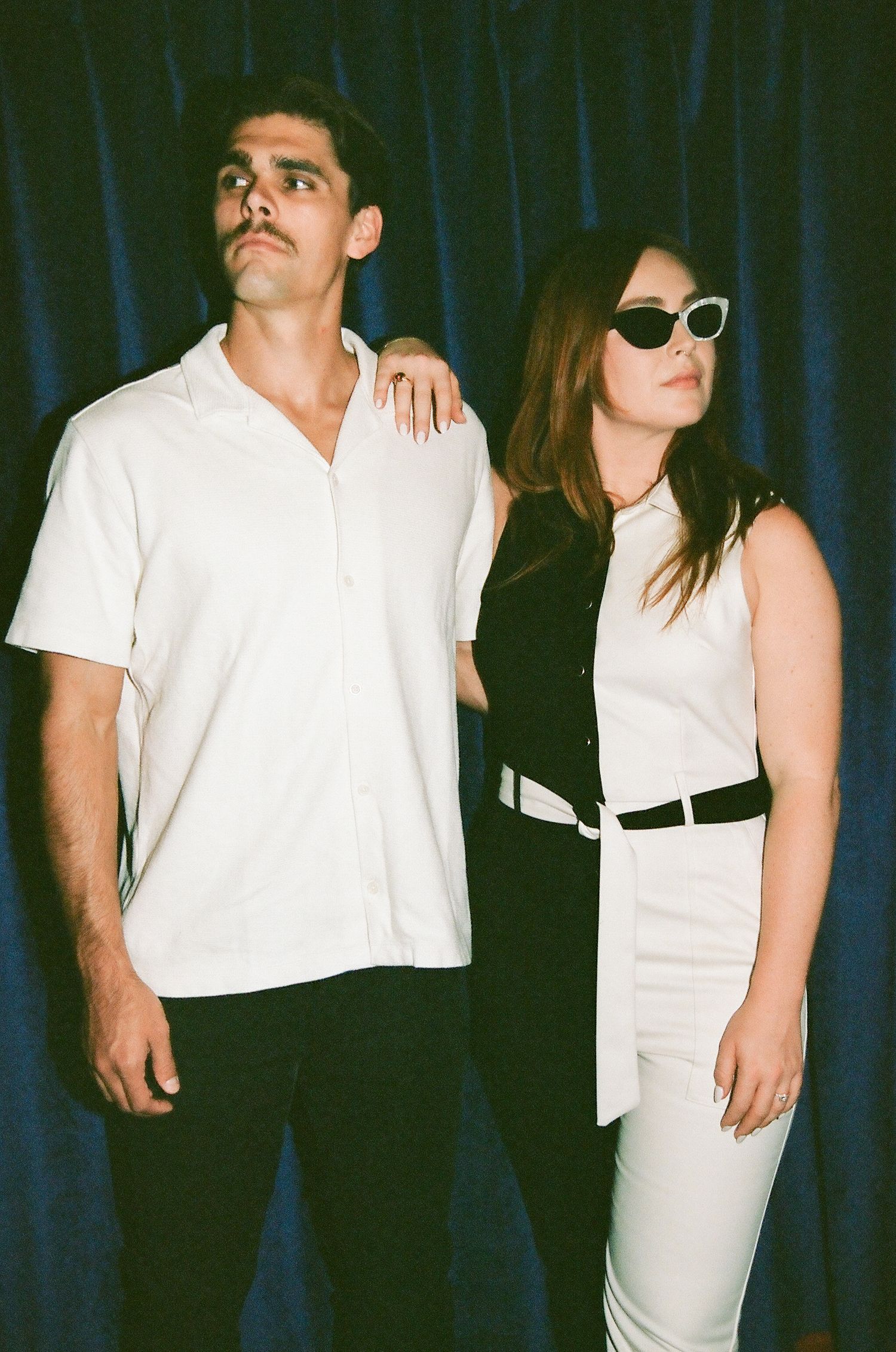 A stylish couple poses against a blue velvet curtain, the woman wearing sunglasses and a black and white outfit.