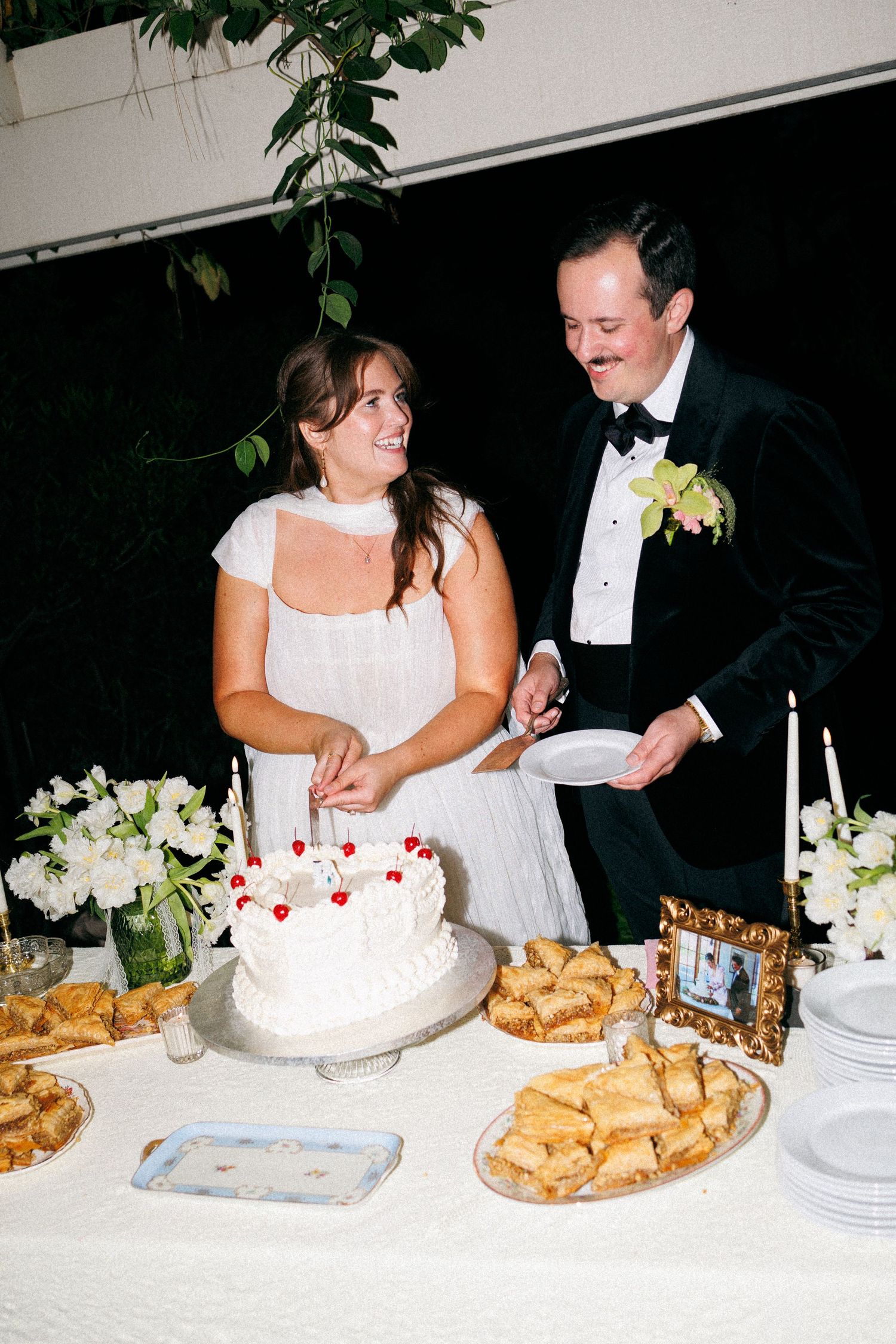 Bride and groom cutting a white wedding cake decorated with cherries at a elegant reception dessert table.