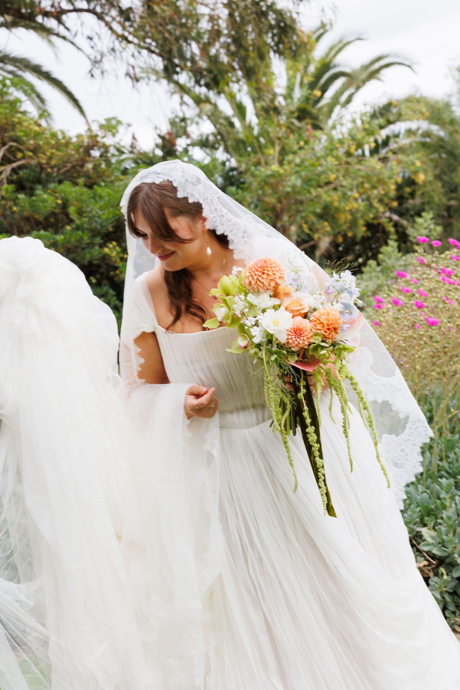 Bride in white gown and lace veil holding colorful peach and green bouquet in garden setting.