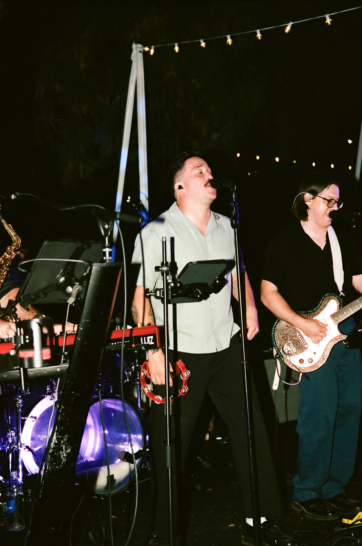 Live band performing outdoors at night, vocalist singing passionately at mic while guitarist plays electric guitar under string lights.