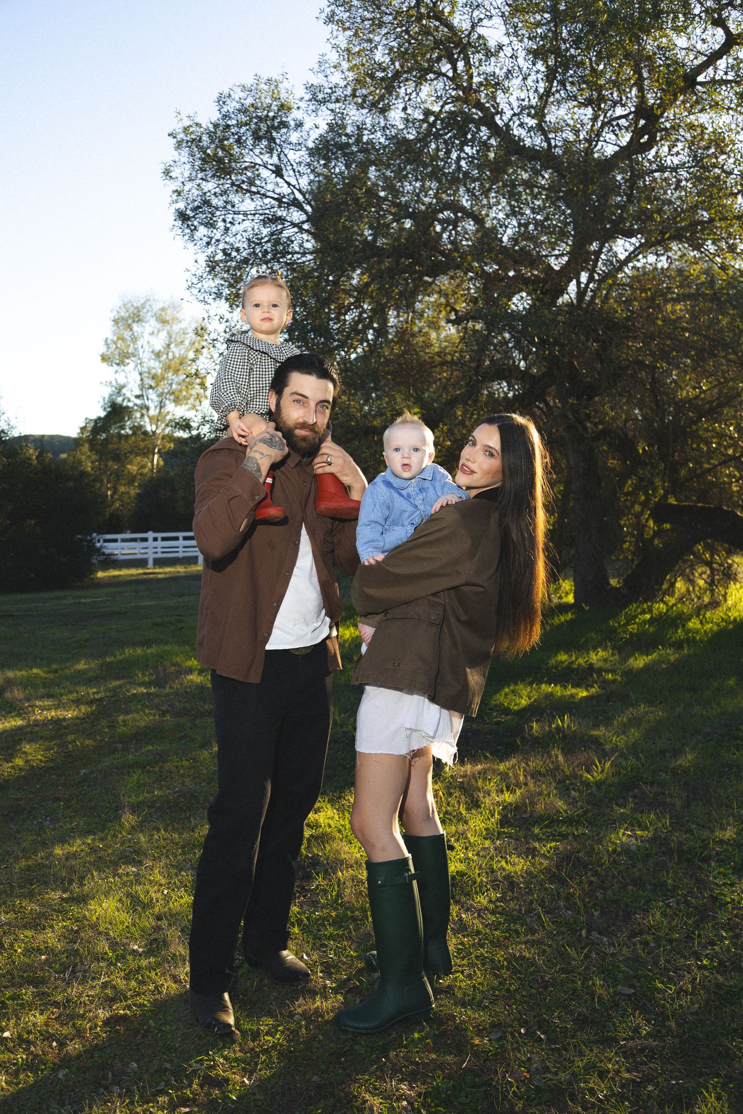 Happy family with bearded father and dark-haired mother holding two babies outdoors in sunny green field.
