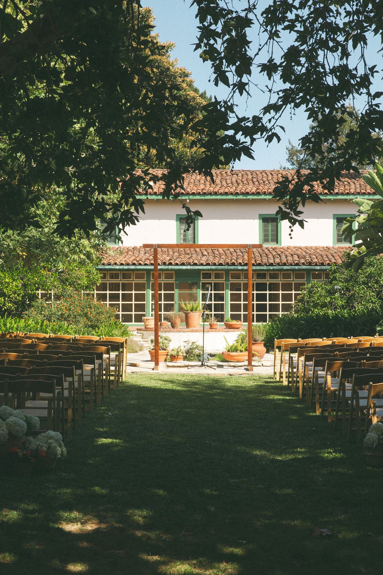 Outdoor wedding ceremony setup with wooden chairs on green lawn facing a rustic white villa with red tile roof.