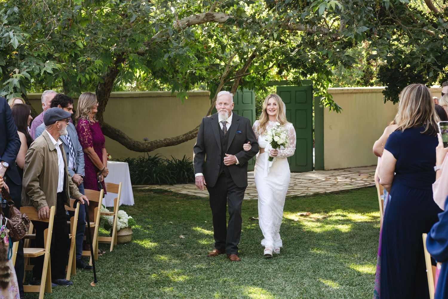 Bride in white gown walks down outdoor aisle with older man, guests seated on chairs, lush garden backdrop.