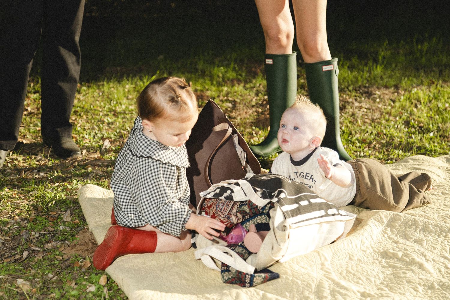 Two young siblings sit together on a blanket outdoors, toddler in red boots and checkered dress with baby.