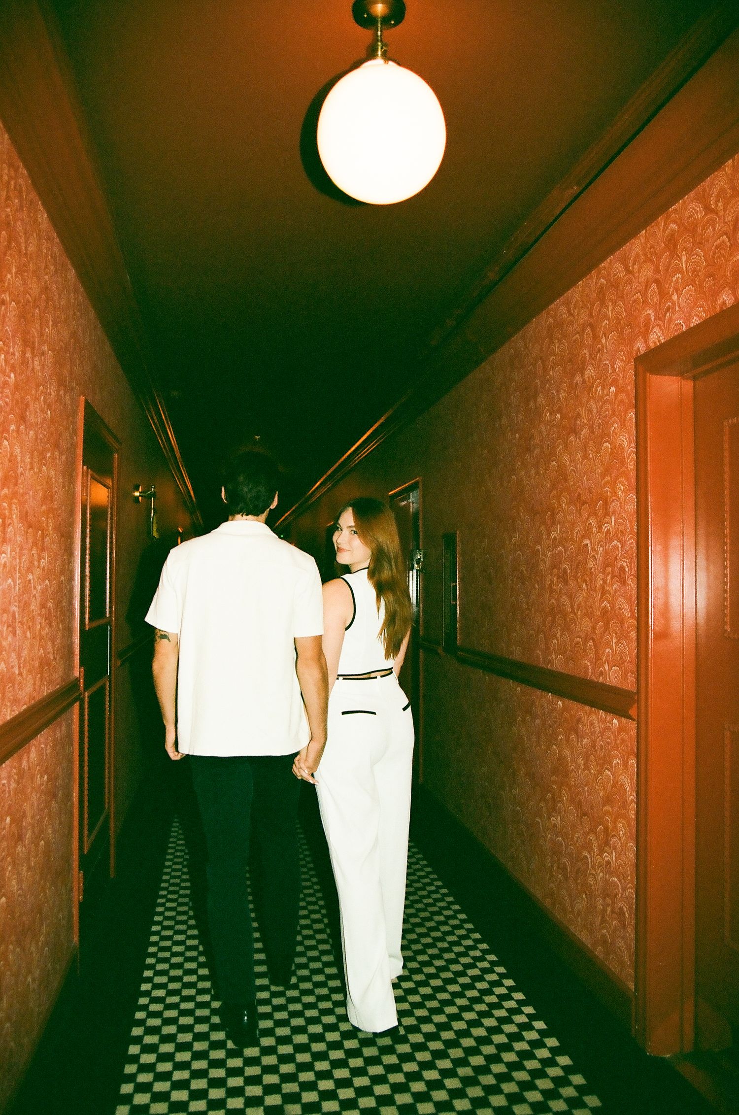 A couple walks hand in hand down a dimly lit hotel hallway with red wallpaper and checkered floors.