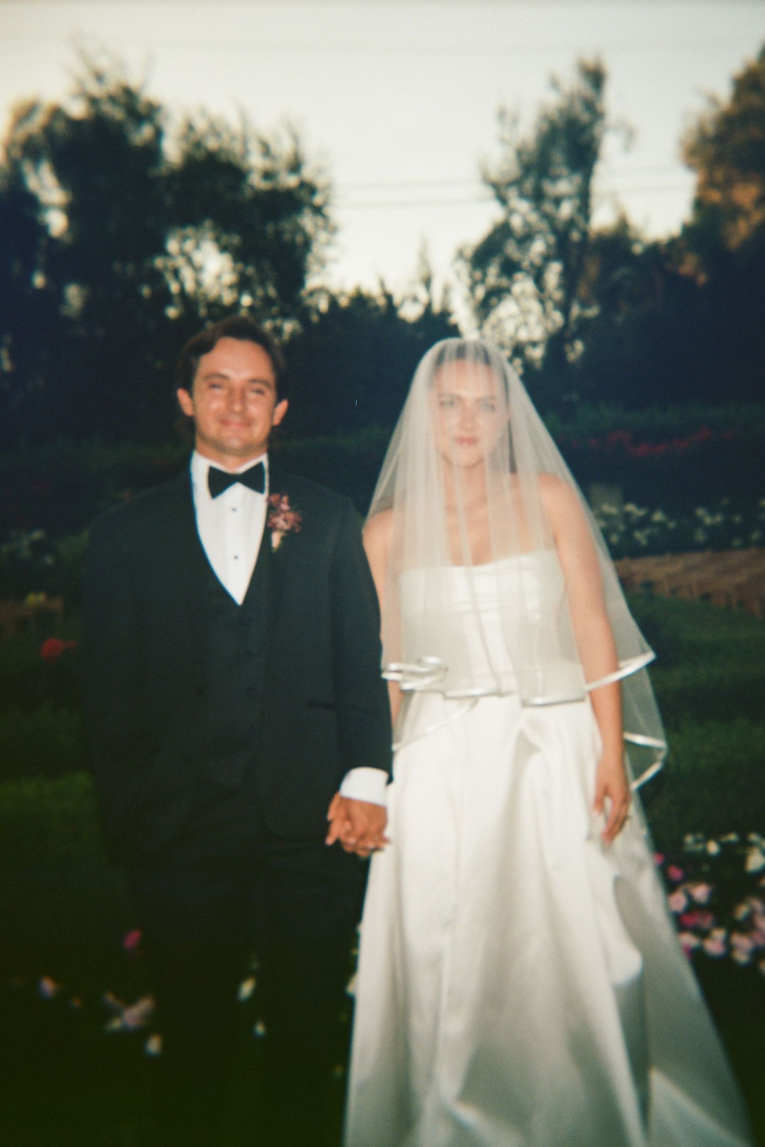 Classic bride and groom pose in garden at dusk, bride in strapless gown with veil, groom in tuxedo.