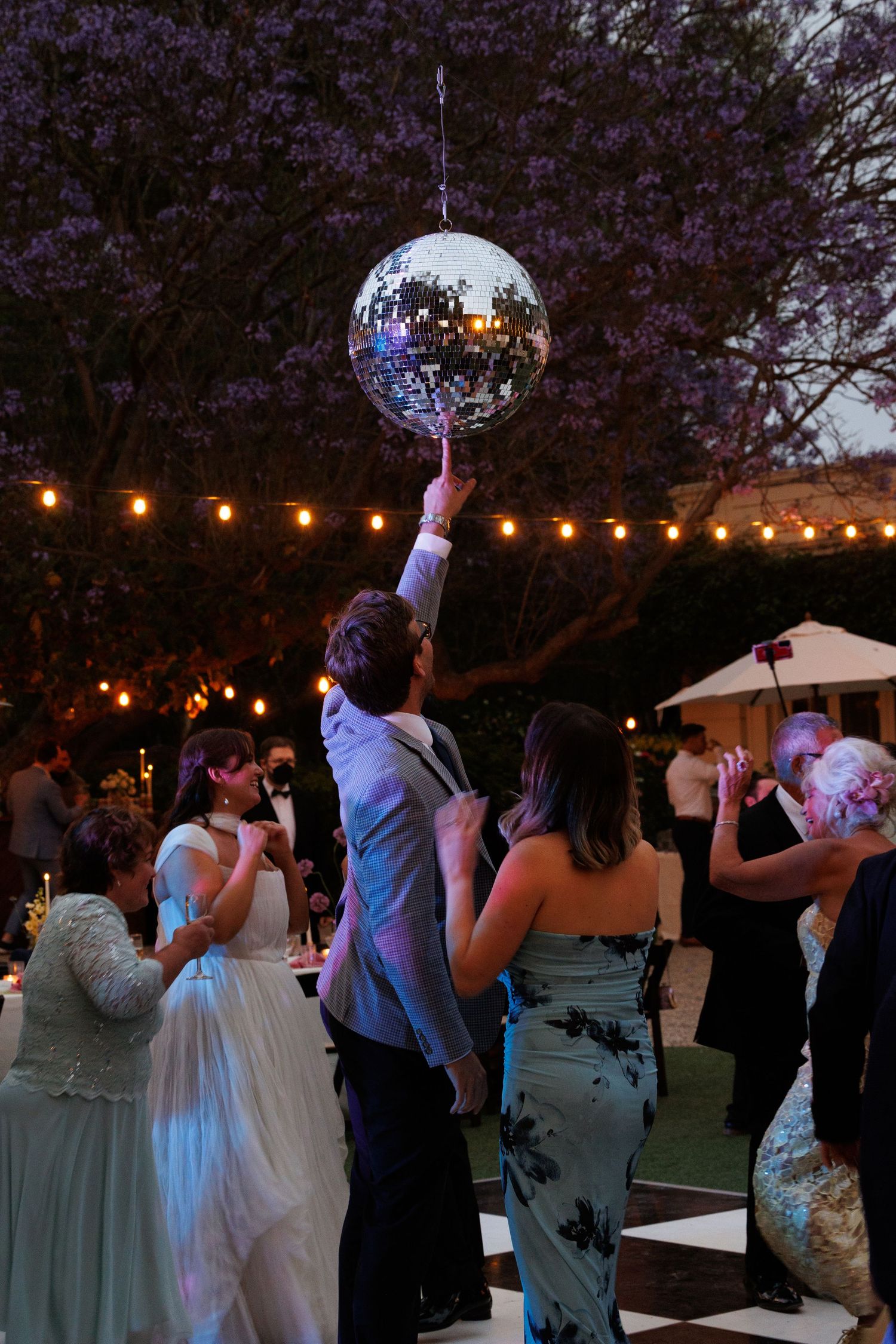 Guest reaches up to touch a disco ball on an outdoor wedding dance floor with string lights and blooming trees.