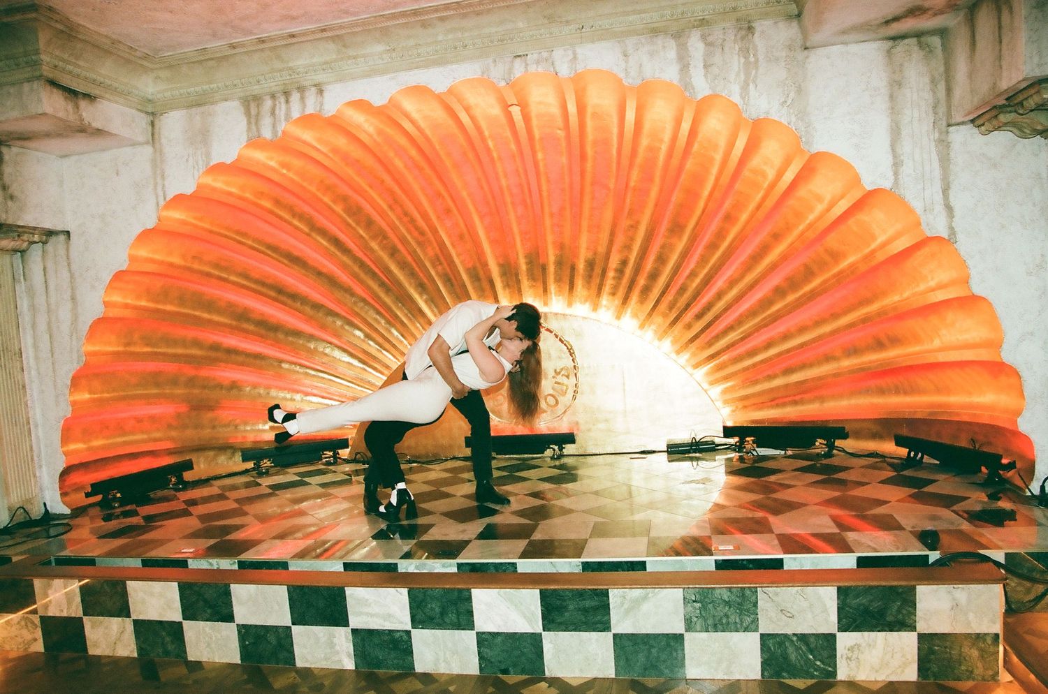 Woman posing dramatically in front of large orange shell-shaped art installation on checkered floor.