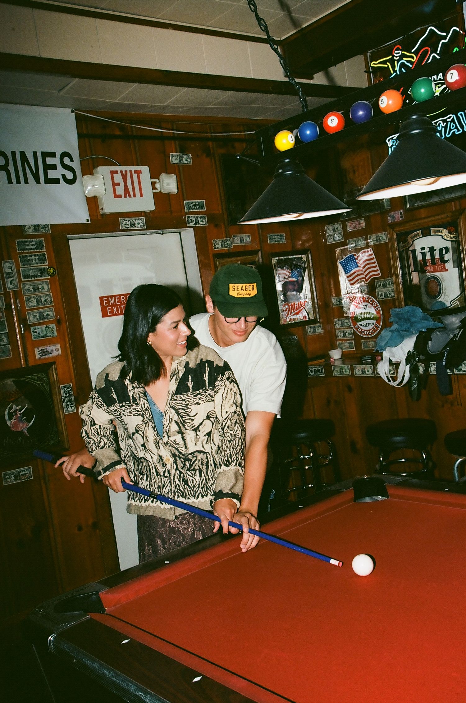 Couple playing pool together at a rustic bar with neon signs and vintage decor.