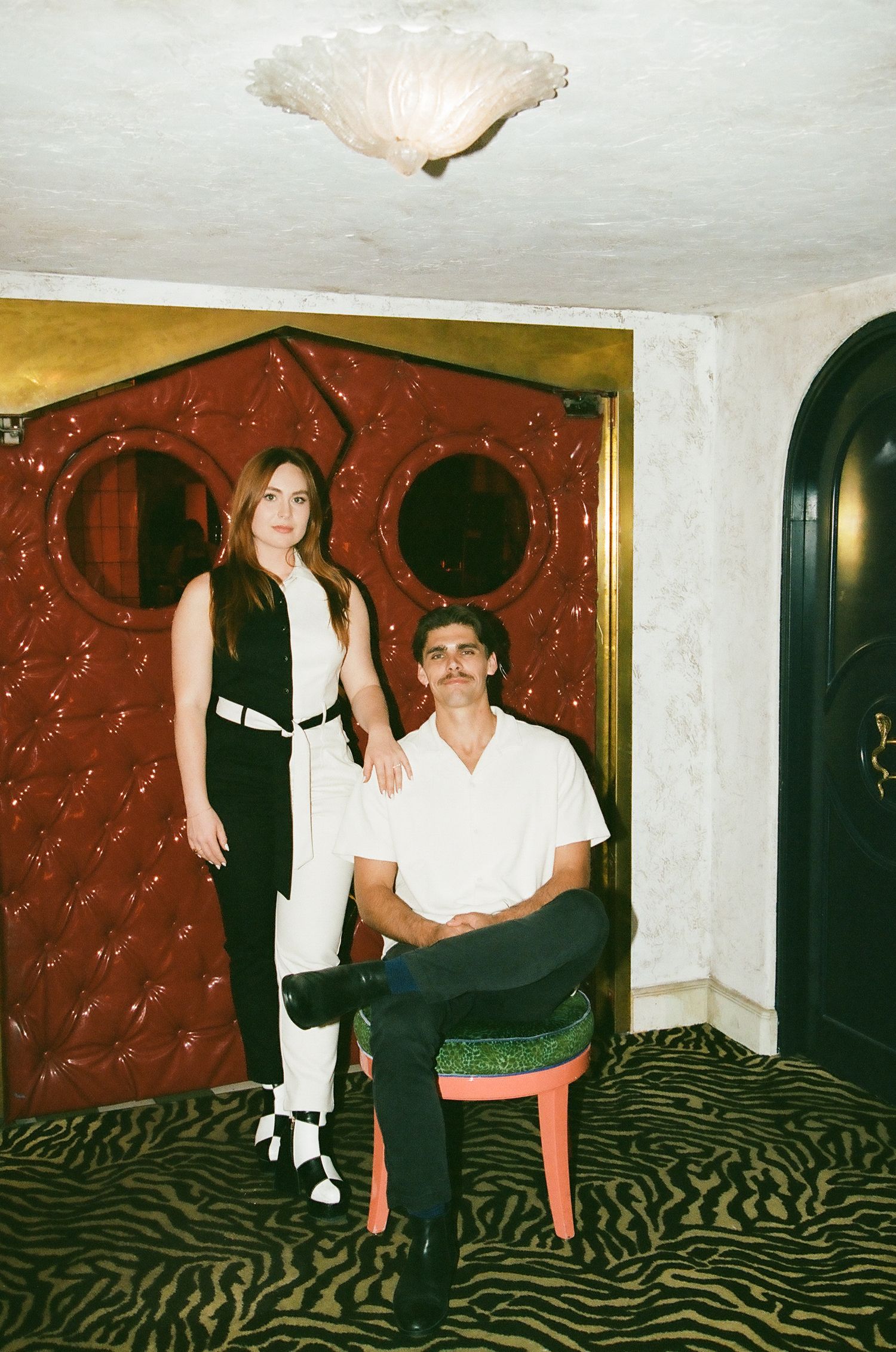 Couple posing in vintage-style room with red tufted wall, woman standing beside seated man.