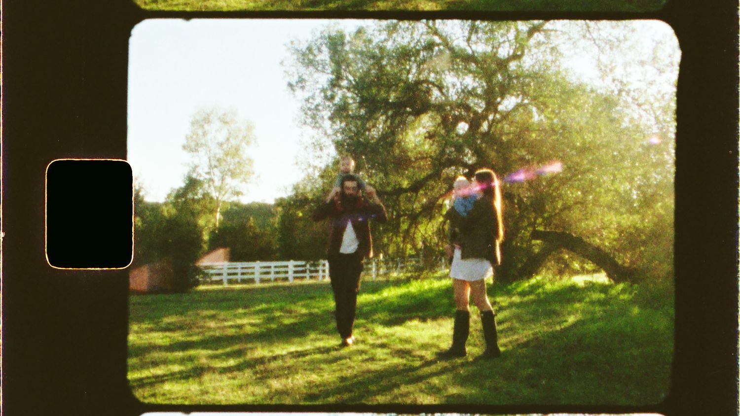 Two people walking in a sunlit green field near a large tree, with a white fence and barn in the background.