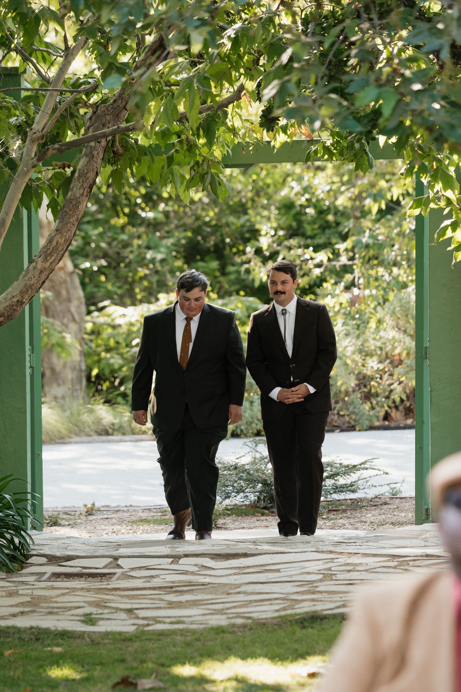 Two men in dark suits walking through a green garden gate on a stone path surrounded by lush trees.