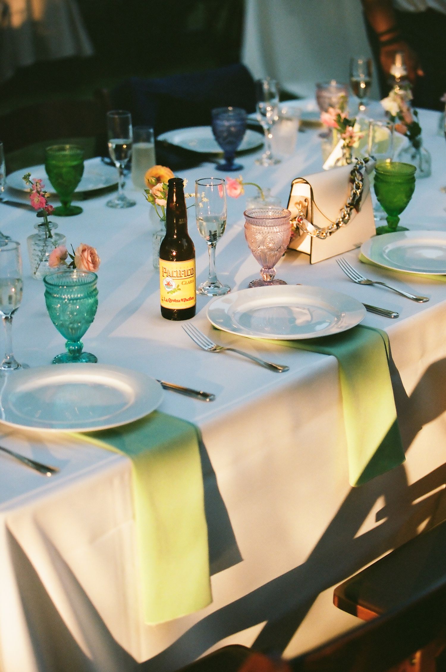 Elegant wedding reception table with colorful glassware, beer bottle centerpiece, white tablecloth, and yellow runner.