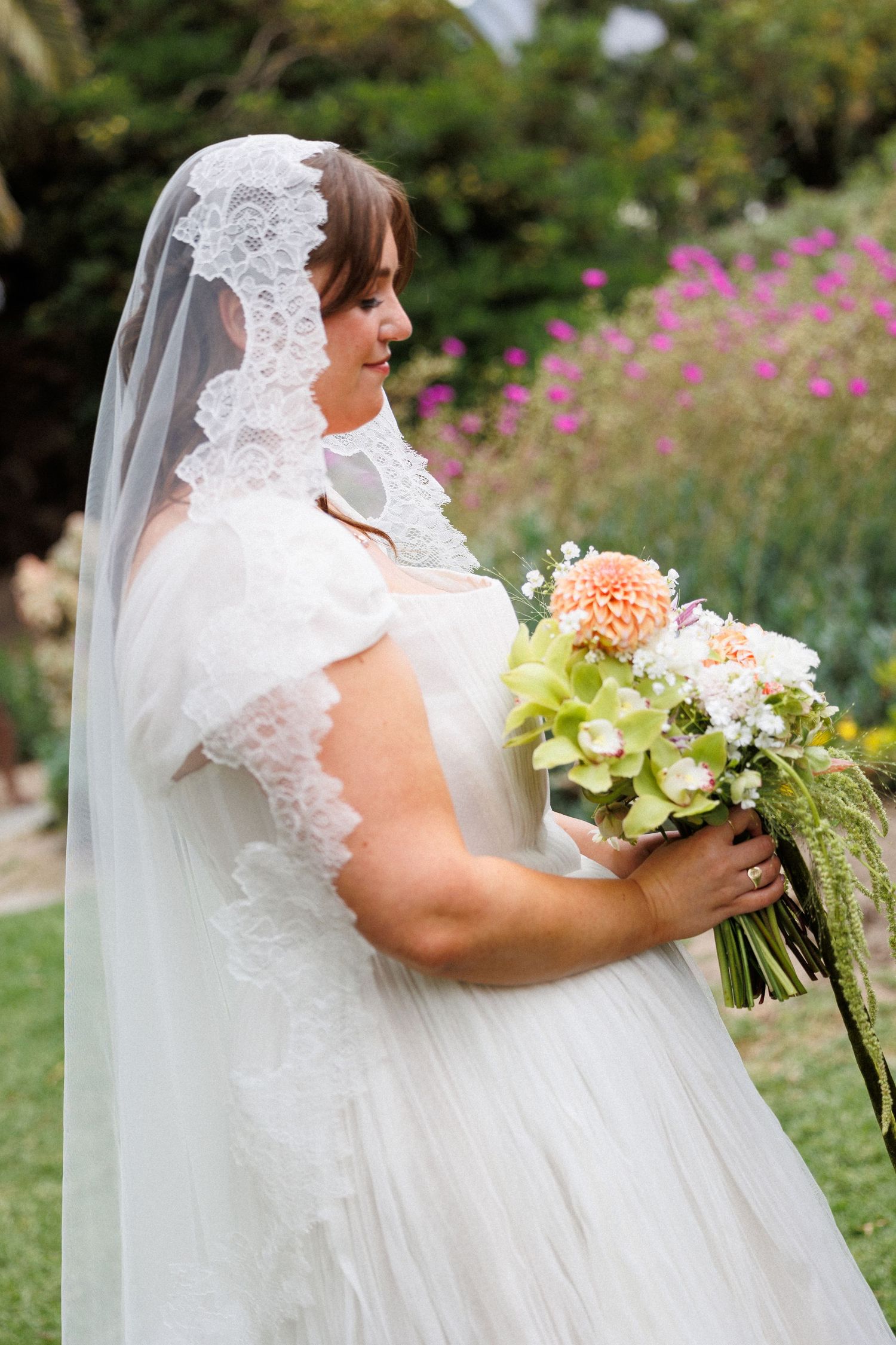 Bride in lace veil and white gown holds elegant bouquet of peach dahlias and green orchids outdoors.