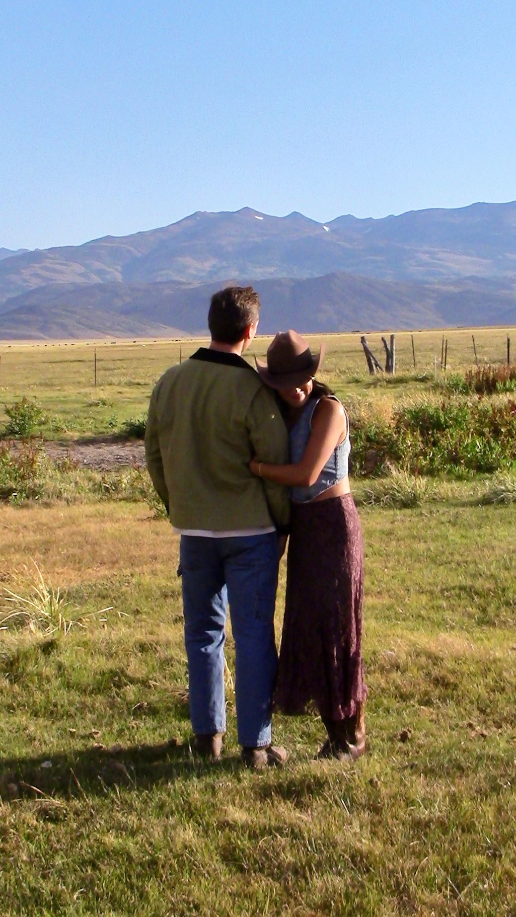 Couple standing together embracing while gazing at a scenic mountain landscape across open fields.