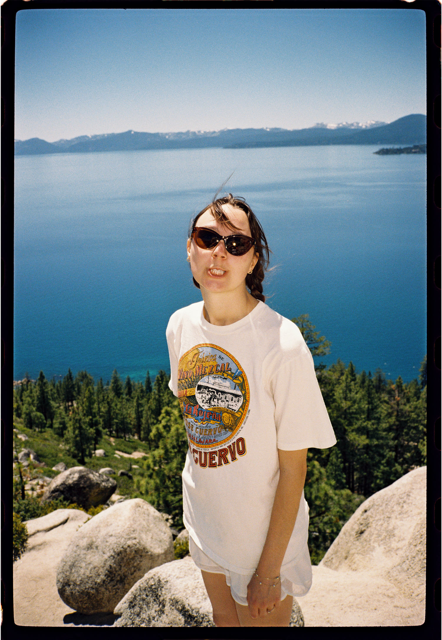 Woman in vintage Cuervo t-shirt poses at scenic Lake Tahoe overlook with deep blue water and mountains behind her.