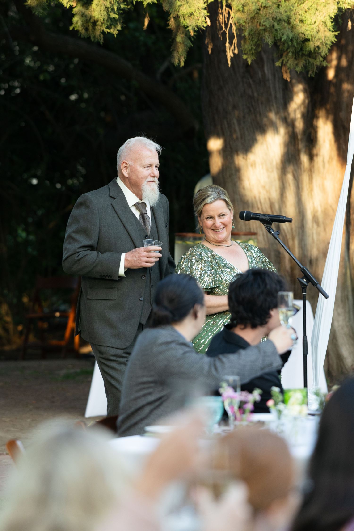 Couple in formal attire giving a toast at an outdoor wedding reception near a microphone stand.