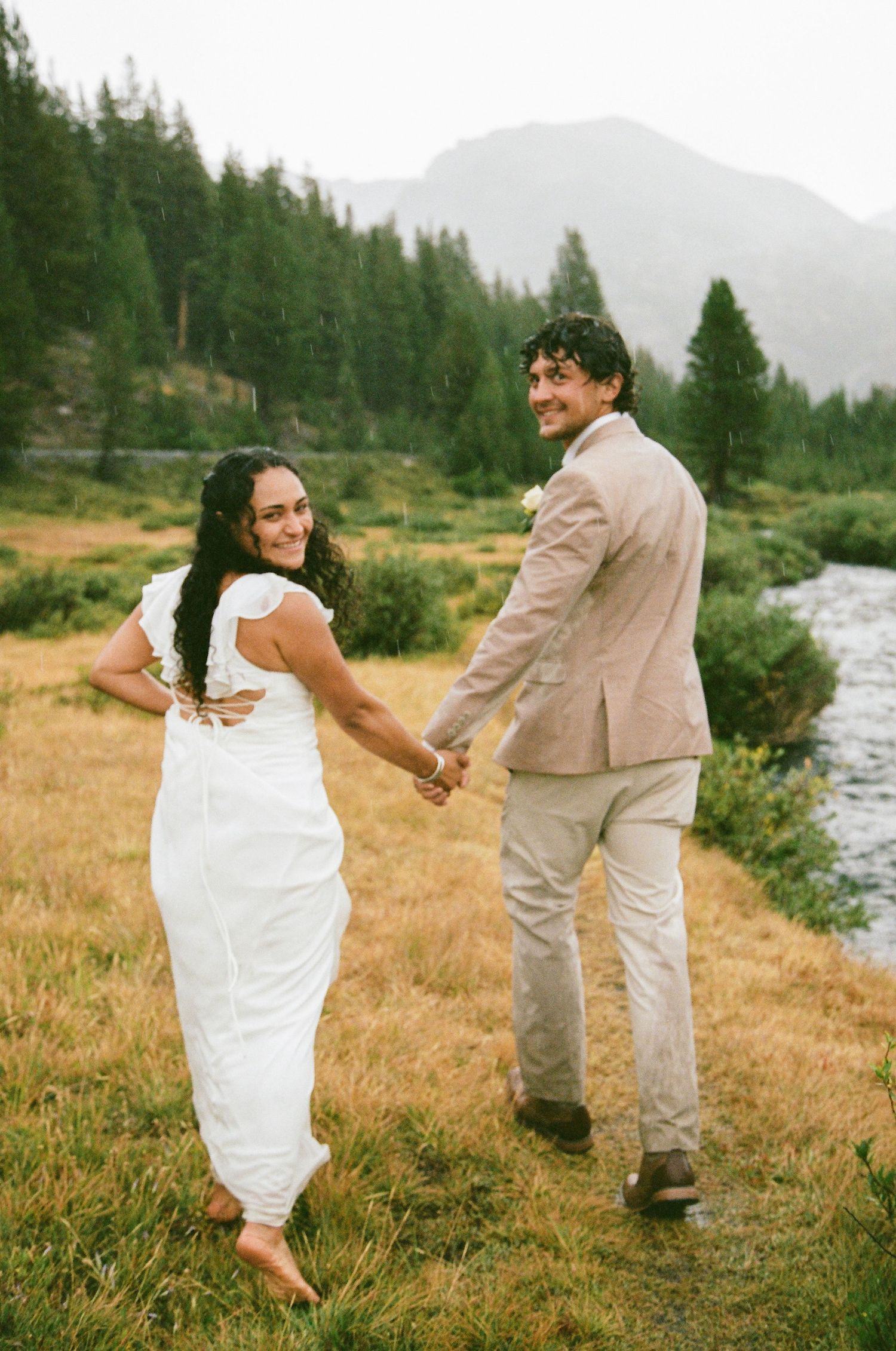Bride in white dress and groom in tan suit hold hands in a mountain meadow by a river, smiling on their wedding day.