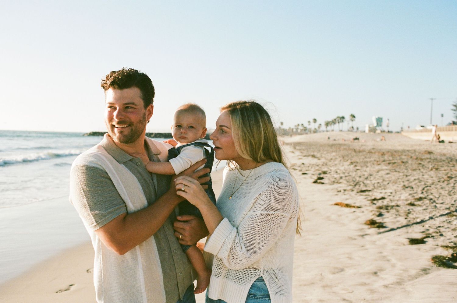 Happy couple holding smiling baby on sunny beach with ocean and palm trees.