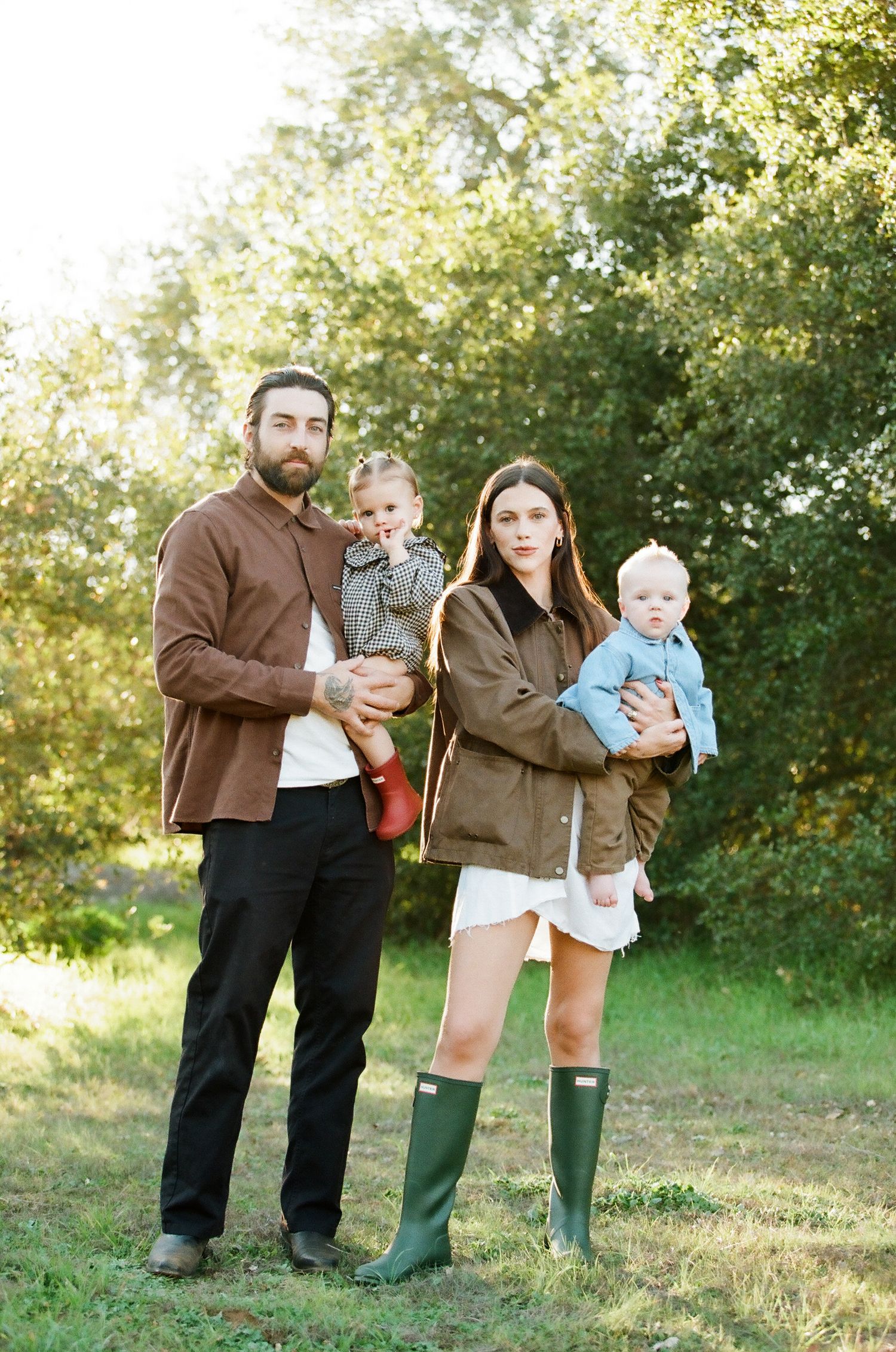 Family of four posing outdoors in autumn, holding two young children in a green setting.