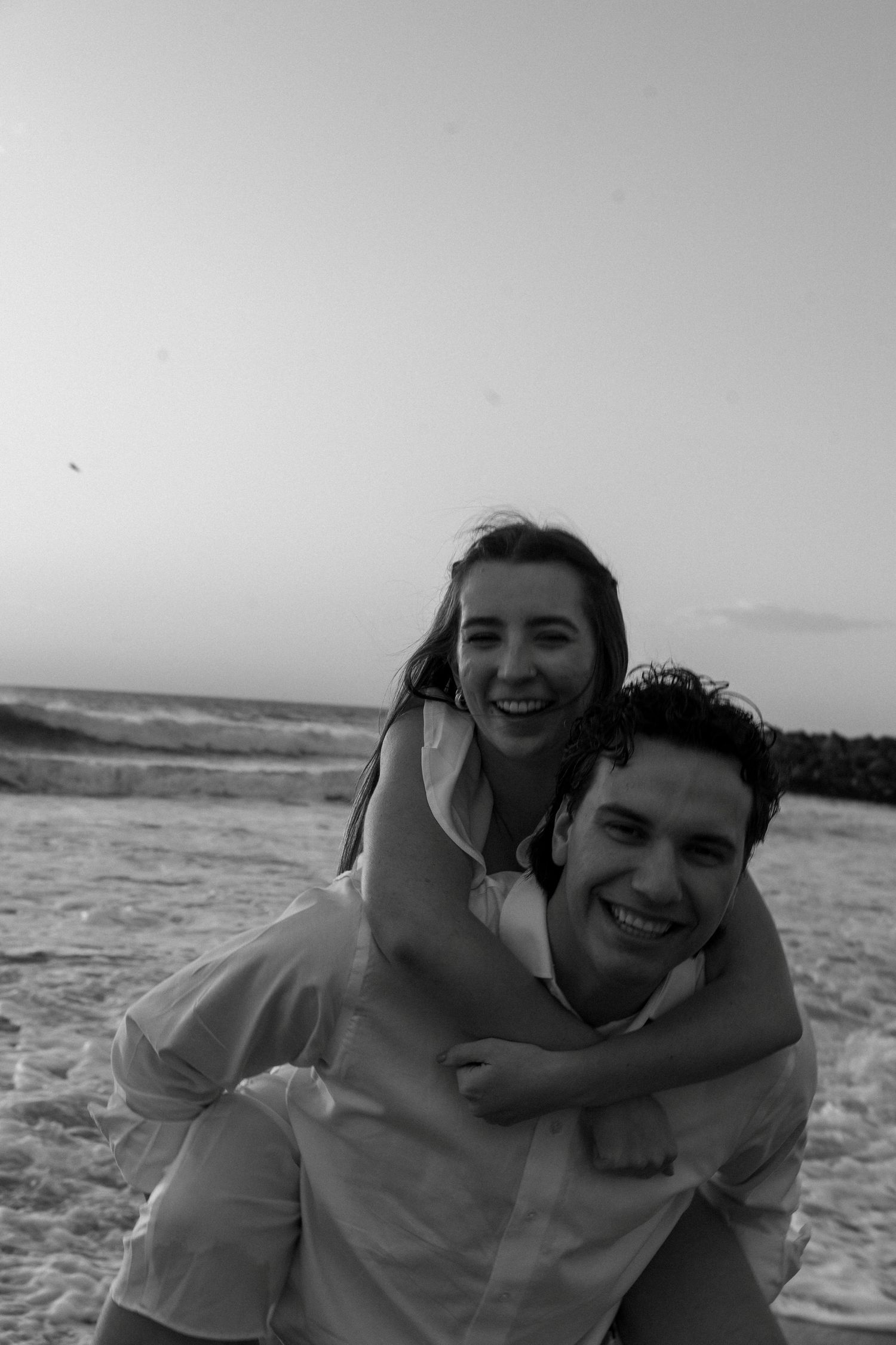 Happy couple in black and white photo, woman riding piggyback on man at beach during sunset.
