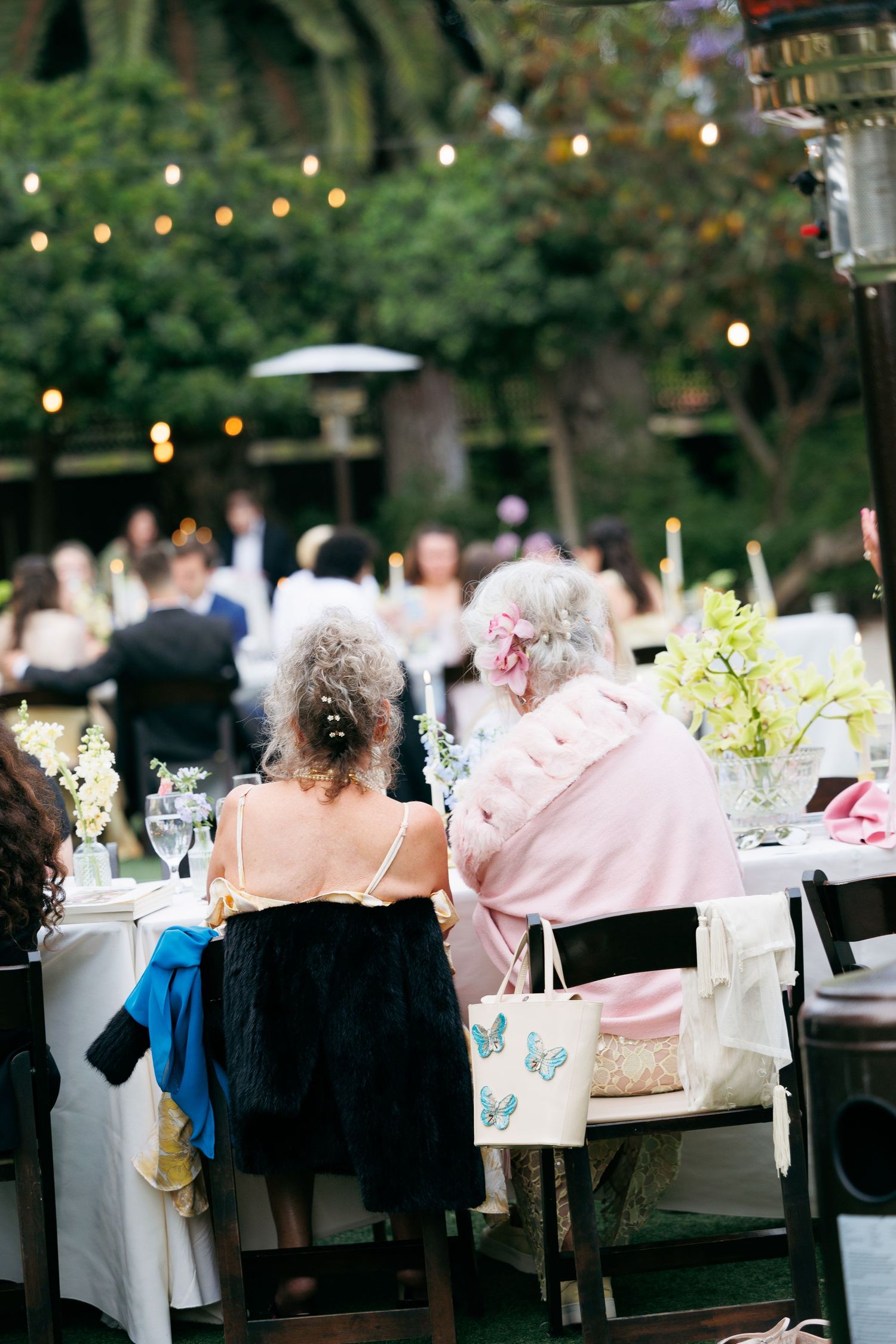 Wedding guests seated at outdoor reception tables with string lights and floral centerpieces at dusk.