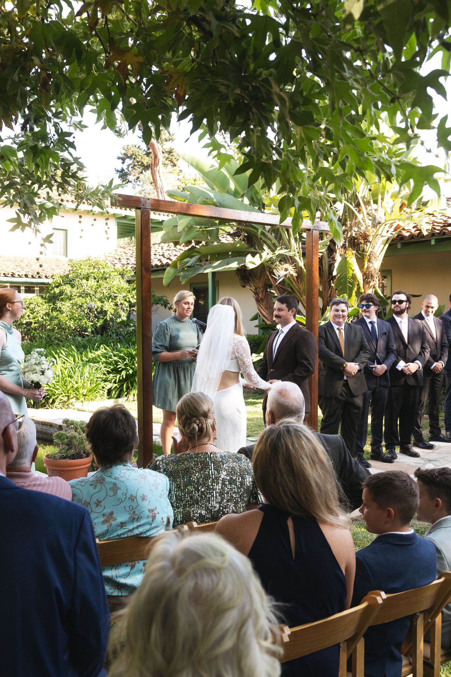 Outdoor wedding ceremony with bride and groom at wooden arch, groomsmen in suits, guests seated on wooden chairs under green trees.