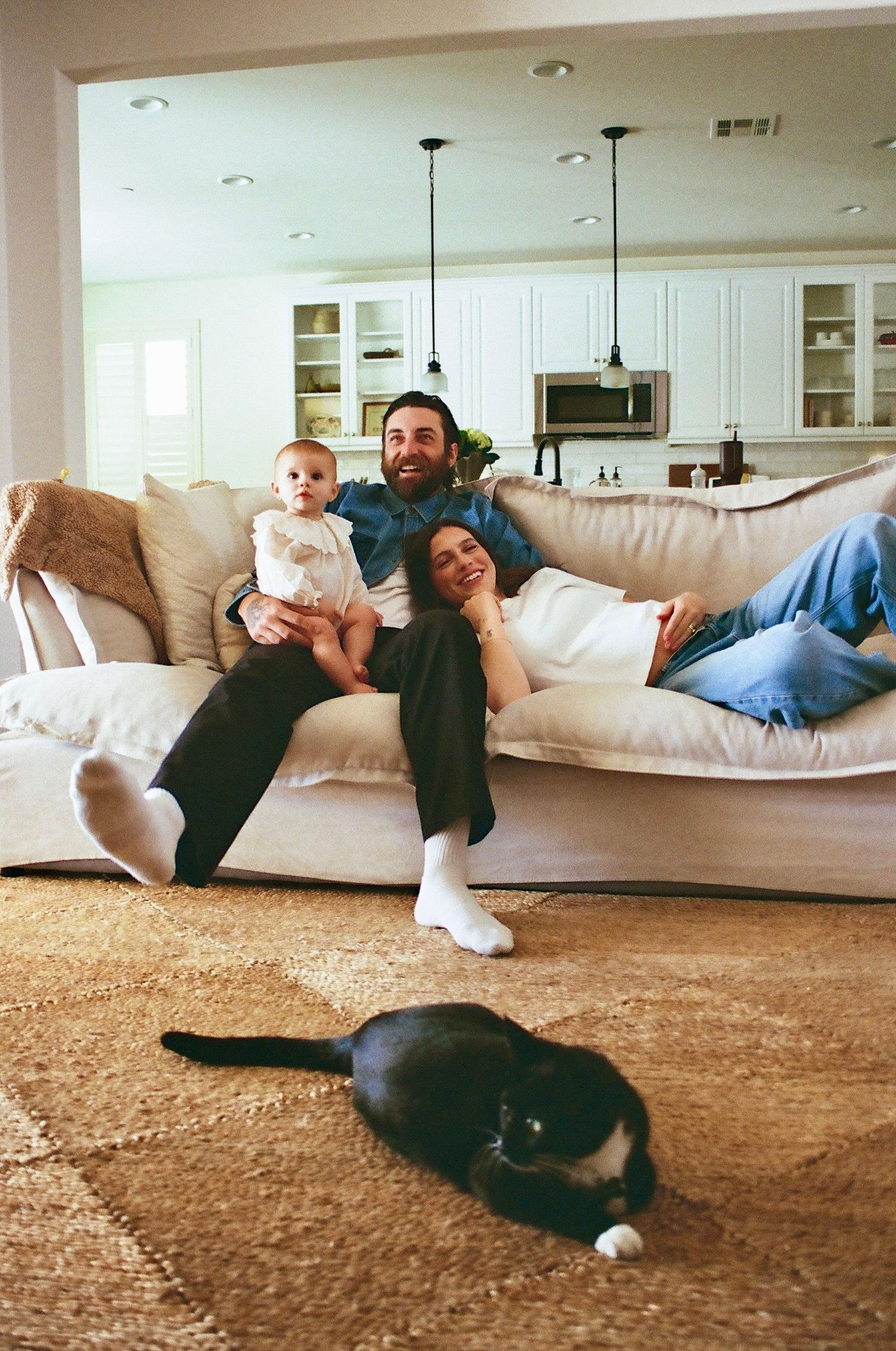 Family relaxing on white sofa with baby and black cat on rug in bright living room.