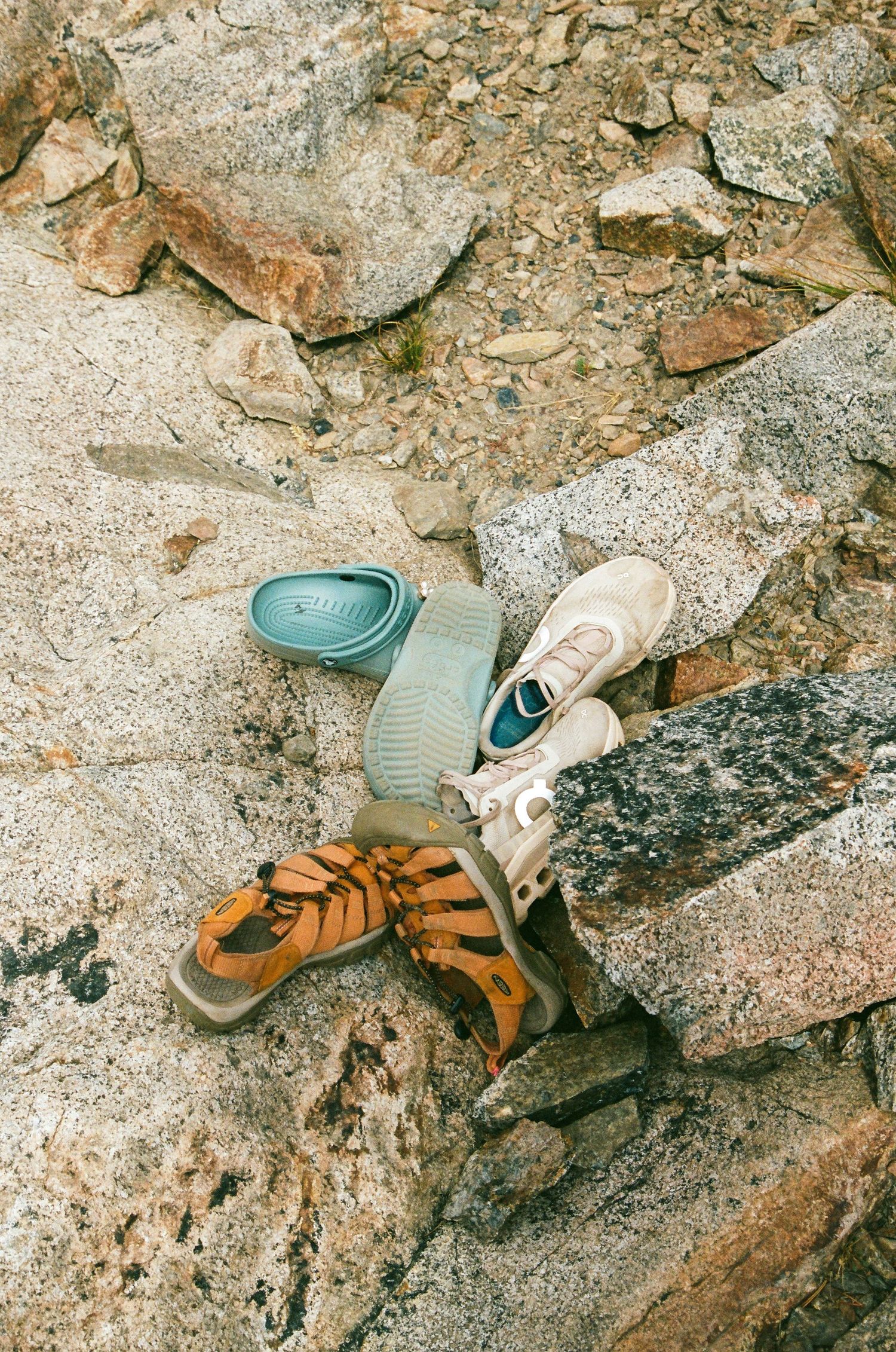 Three pairs of shoes on rocky terrain: teal slides, beige sneakers, and orange sandals arranged outdoors.