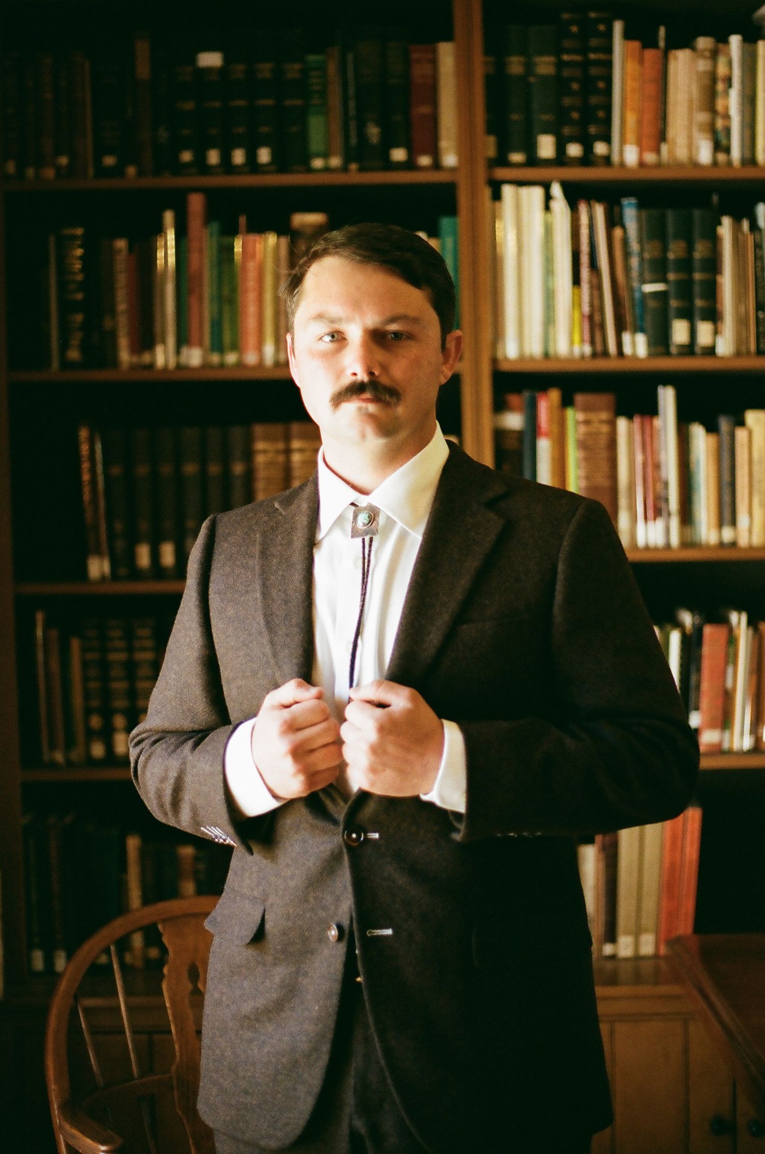 Man with mustache in dark blazer and bolo tie standing in front of bookshelf in a library.