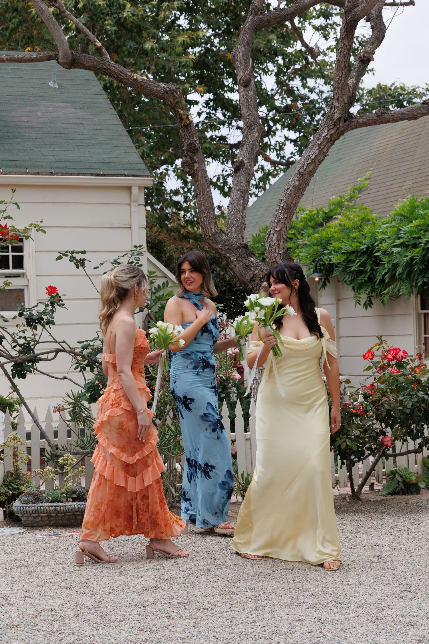 Three bridesmaids in colorful dresses holding white flowers outside a white cottage.