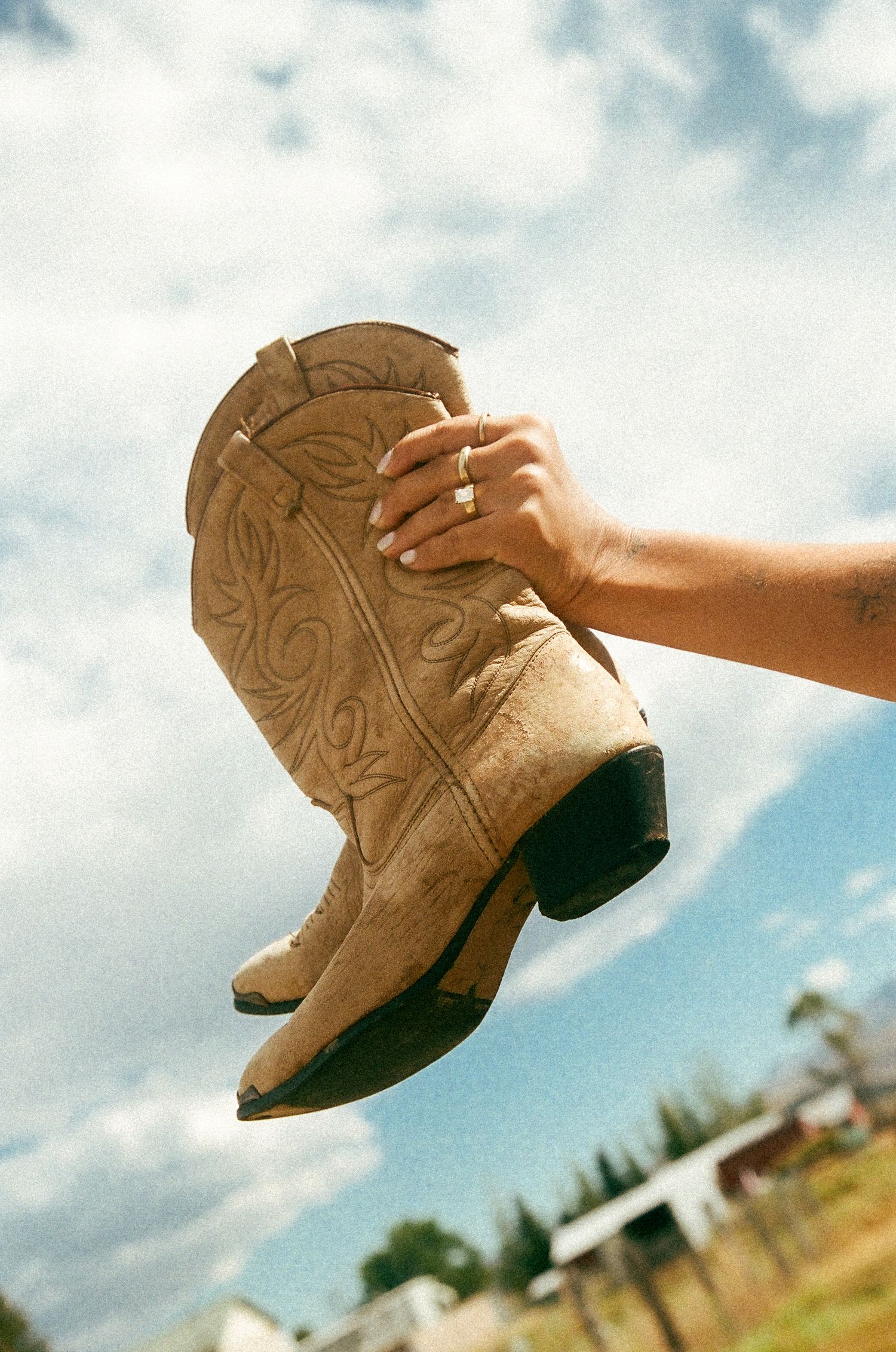 A hand holds tan leather cowboy boots with decorative stitching against a cloudy sky.