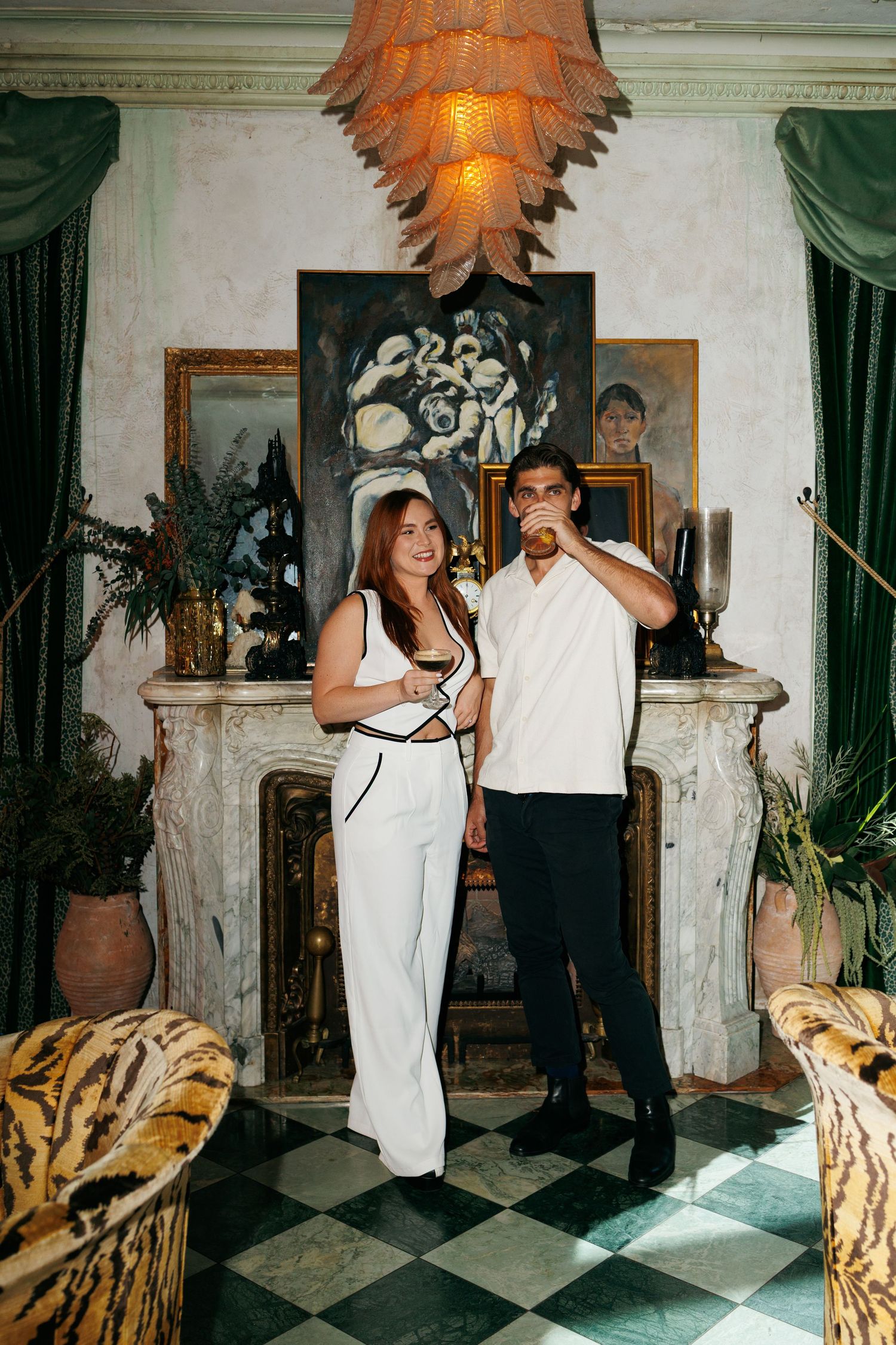 Couple posing in elegant room with fireplace, artwork, and chandelier, woman in white jumpsuit holding wine glass.