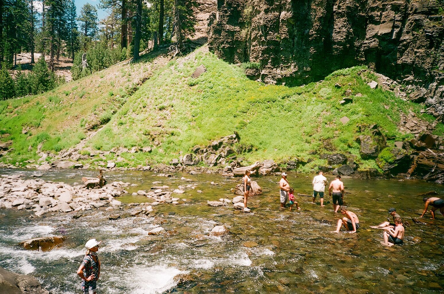 People wading in a rocky mountain river surrounded by lush green hills and pine trees.