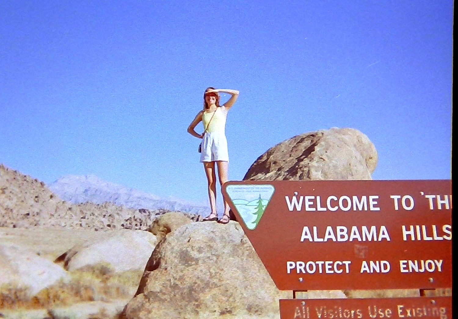 Woman standing on a boulder at the Welcome to Alabama Hills sign in a desert landscape.
