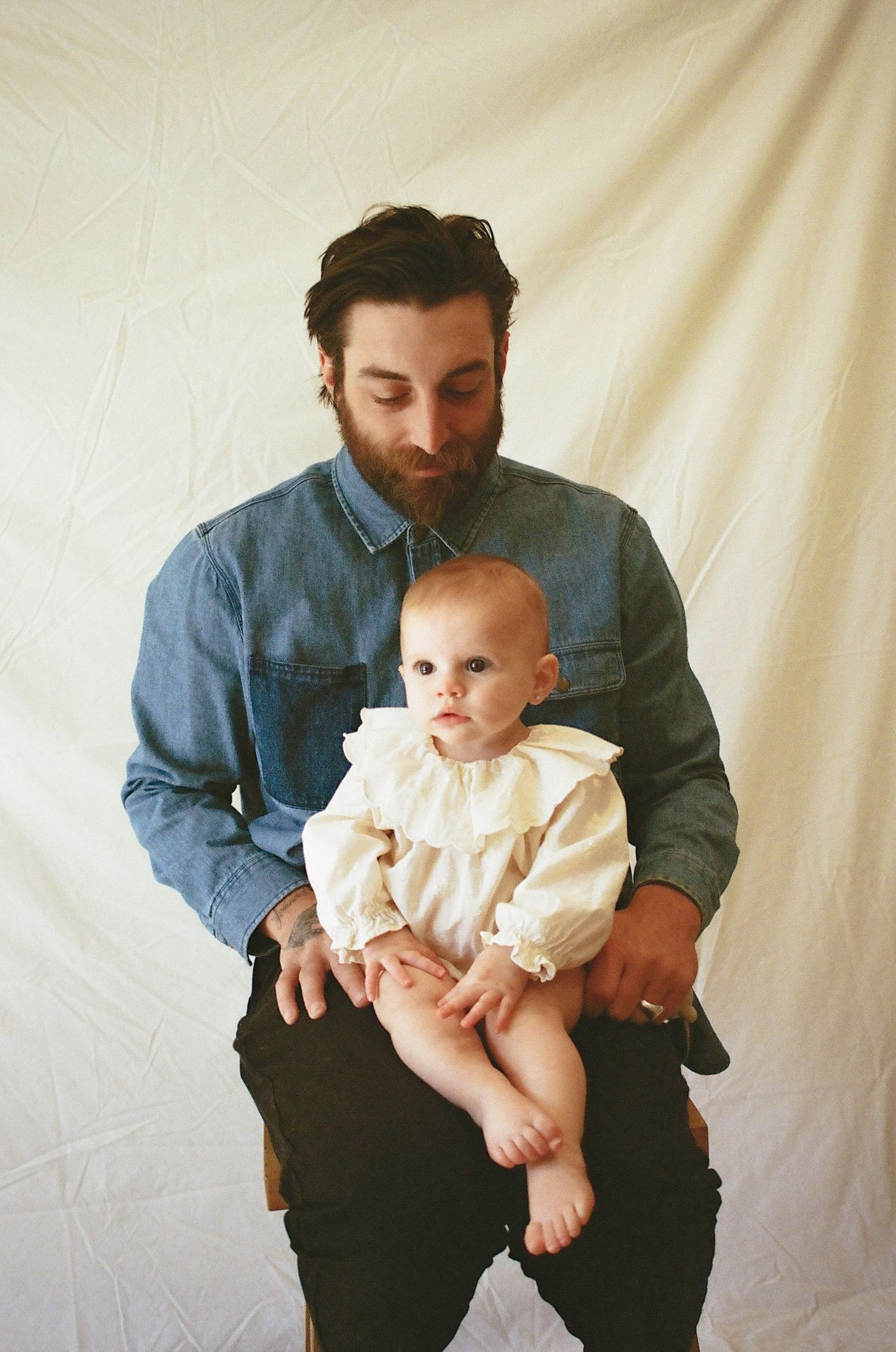 Bearded father in denim shirt holding baby in ruffled outfit against white backdrop.