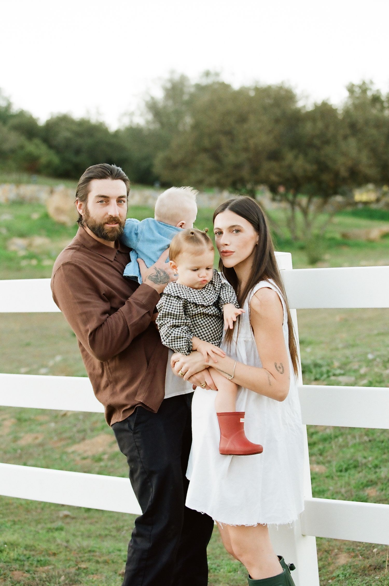 A young family with two toddlers poses by a white fence on a green farm, parents dressed casually in brown and white.
