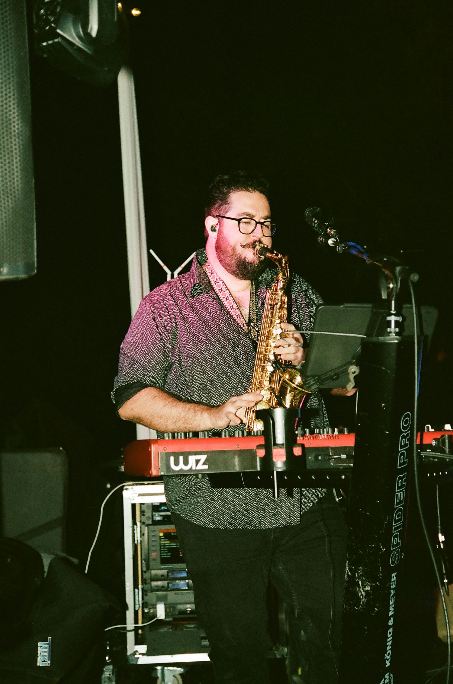 Man playing saxophone on stage with keyboard and microphone in dimly lit venue.