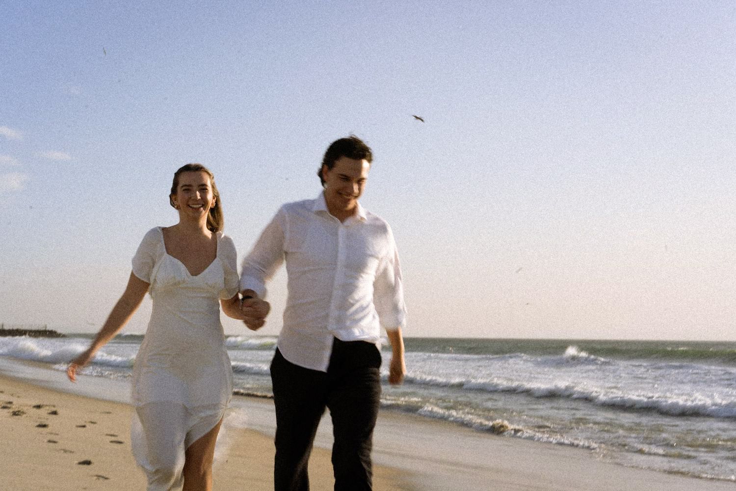 Happy couple holding hands and walking along a sunny beach with ocean waves behind them.