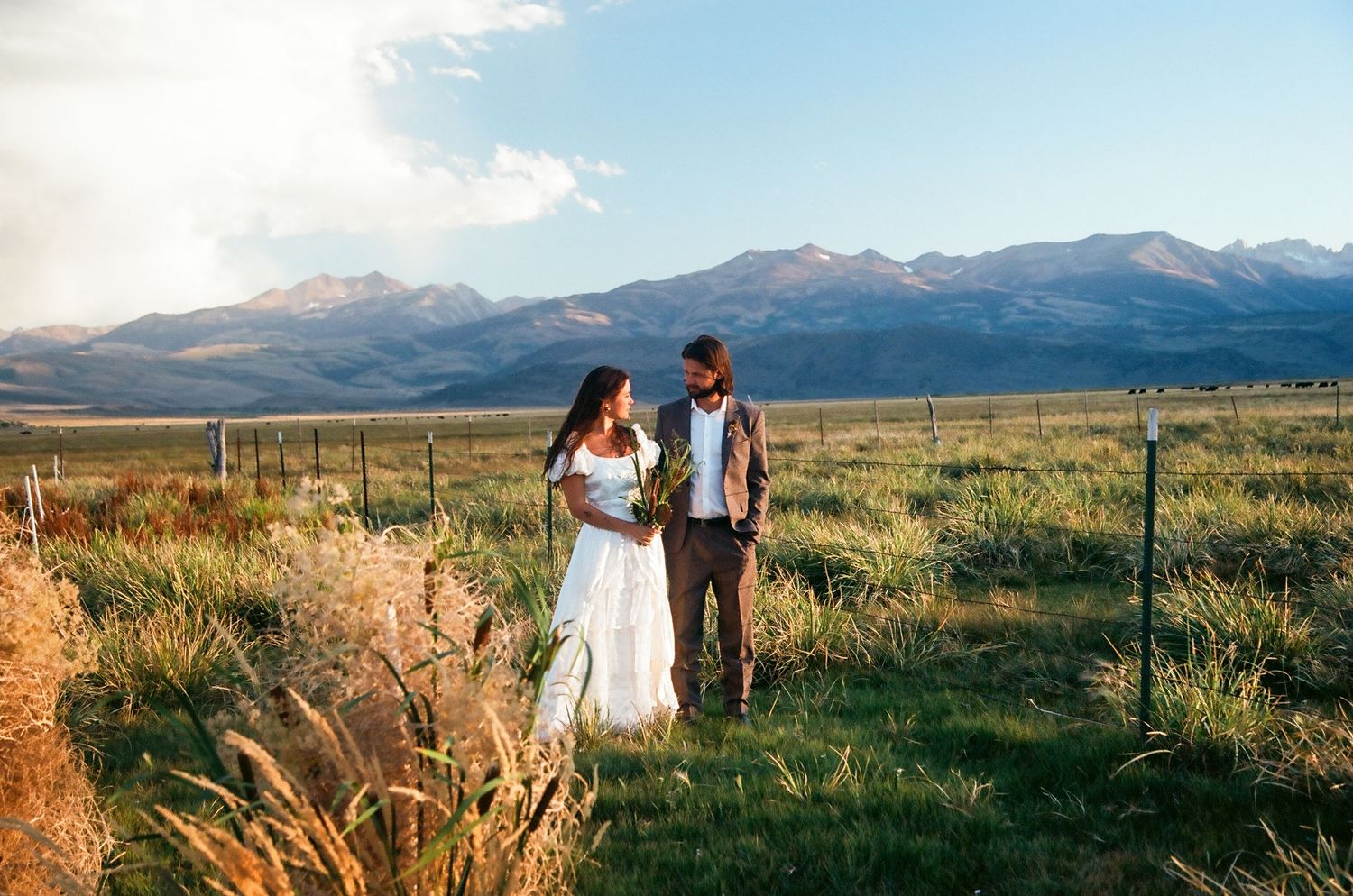 Bride and groom holding hands in scenic mountain meadow at golden hour sunset.