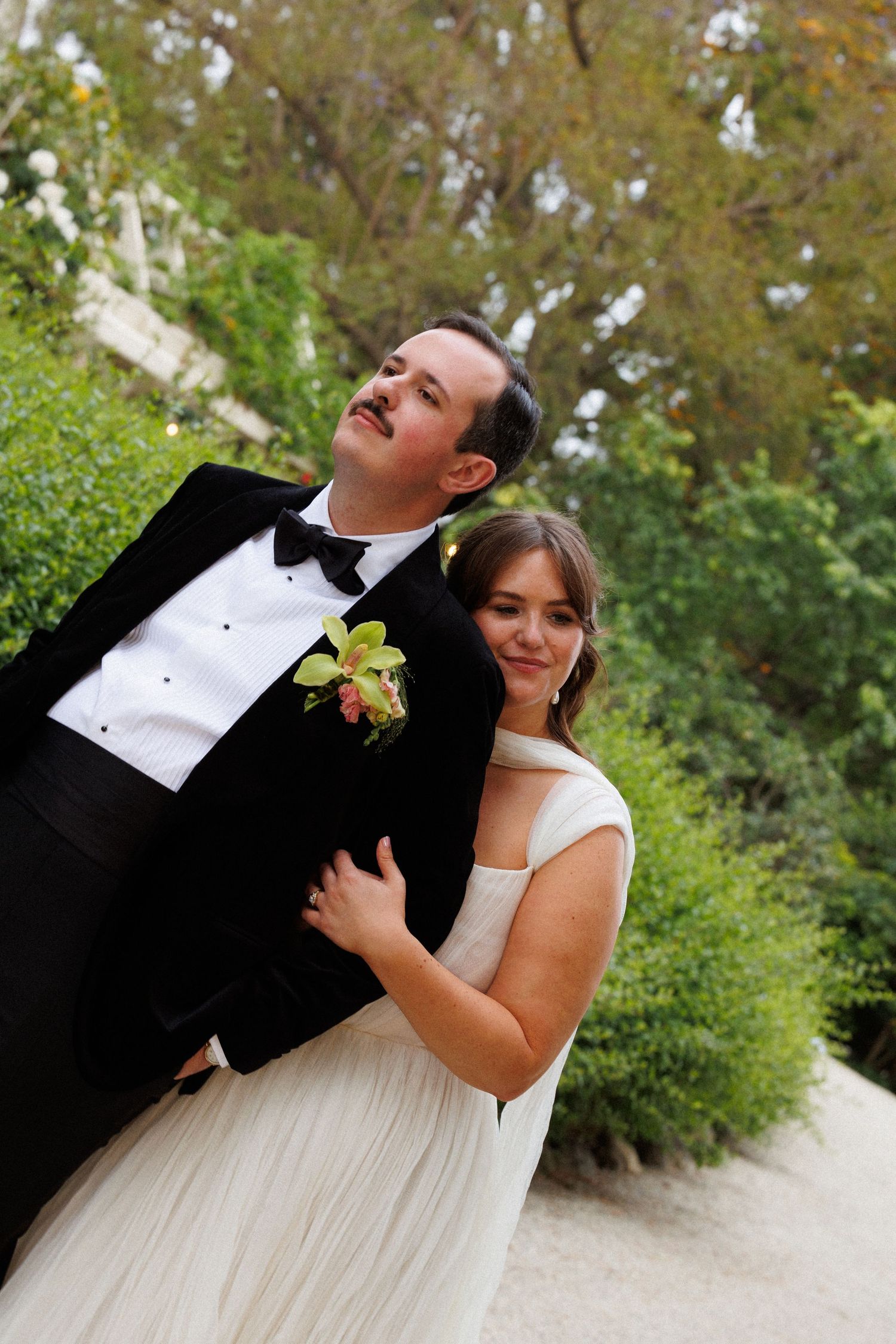 A bride in a white gown embraces a groom in a black tuxedo with a green boutonniere outdoors among lush greenery.