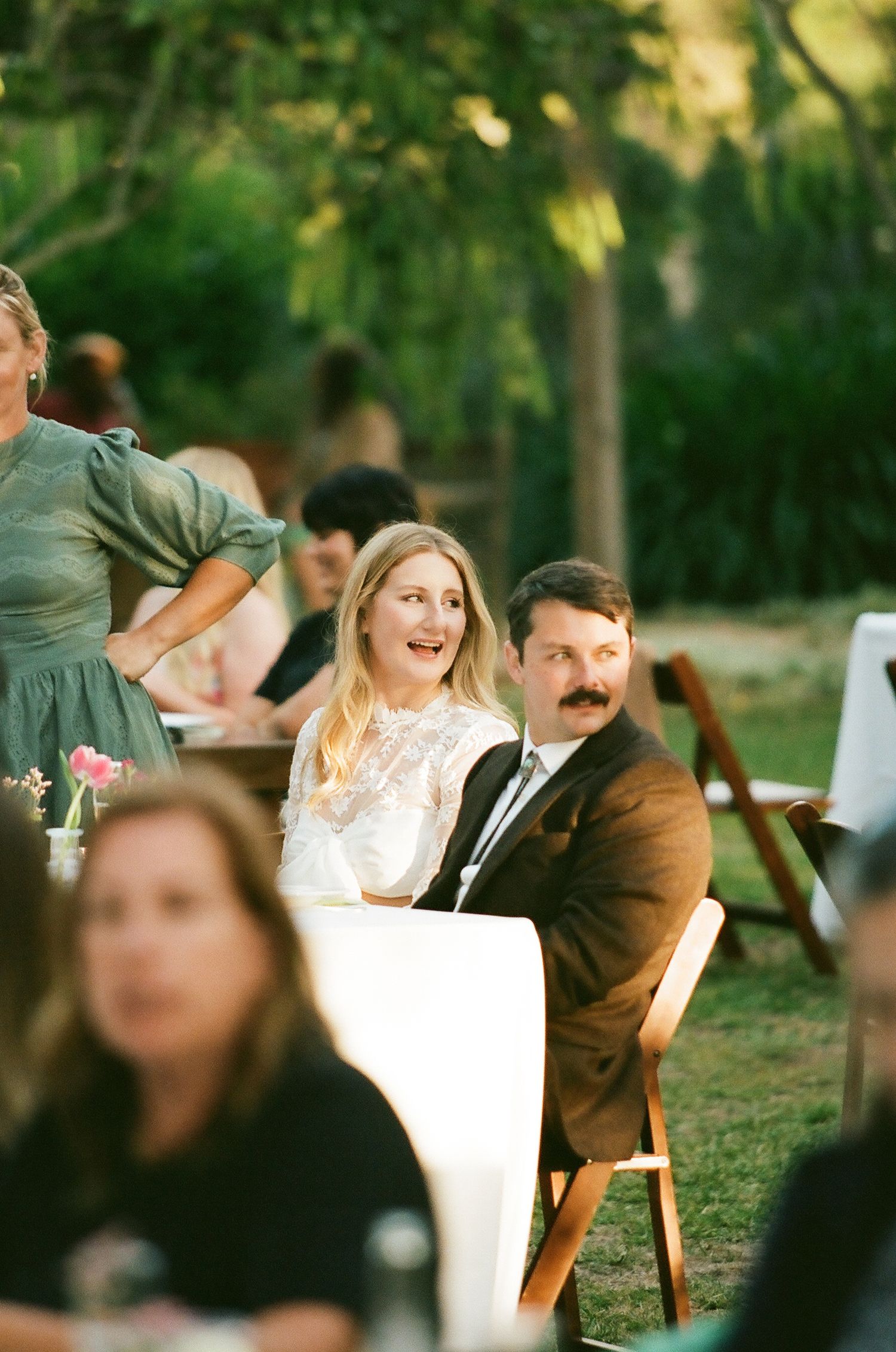Bride and groom seated outdoors at wedding reception table, smiling, surrounded by guests in garden setting.