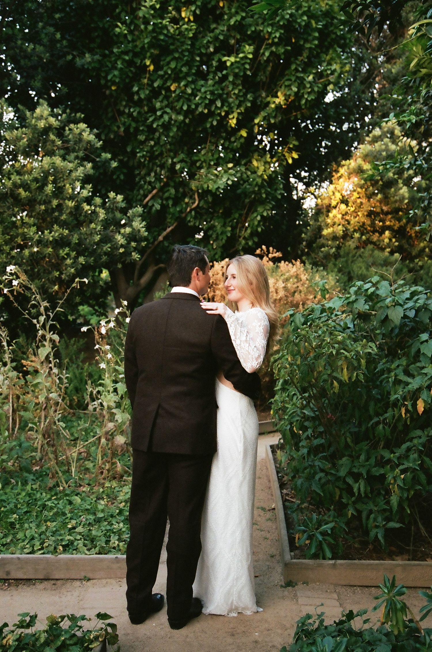 A bride and groom share a romantic moment in a lush garden, she wearing a white lace gown and he a dark suit.